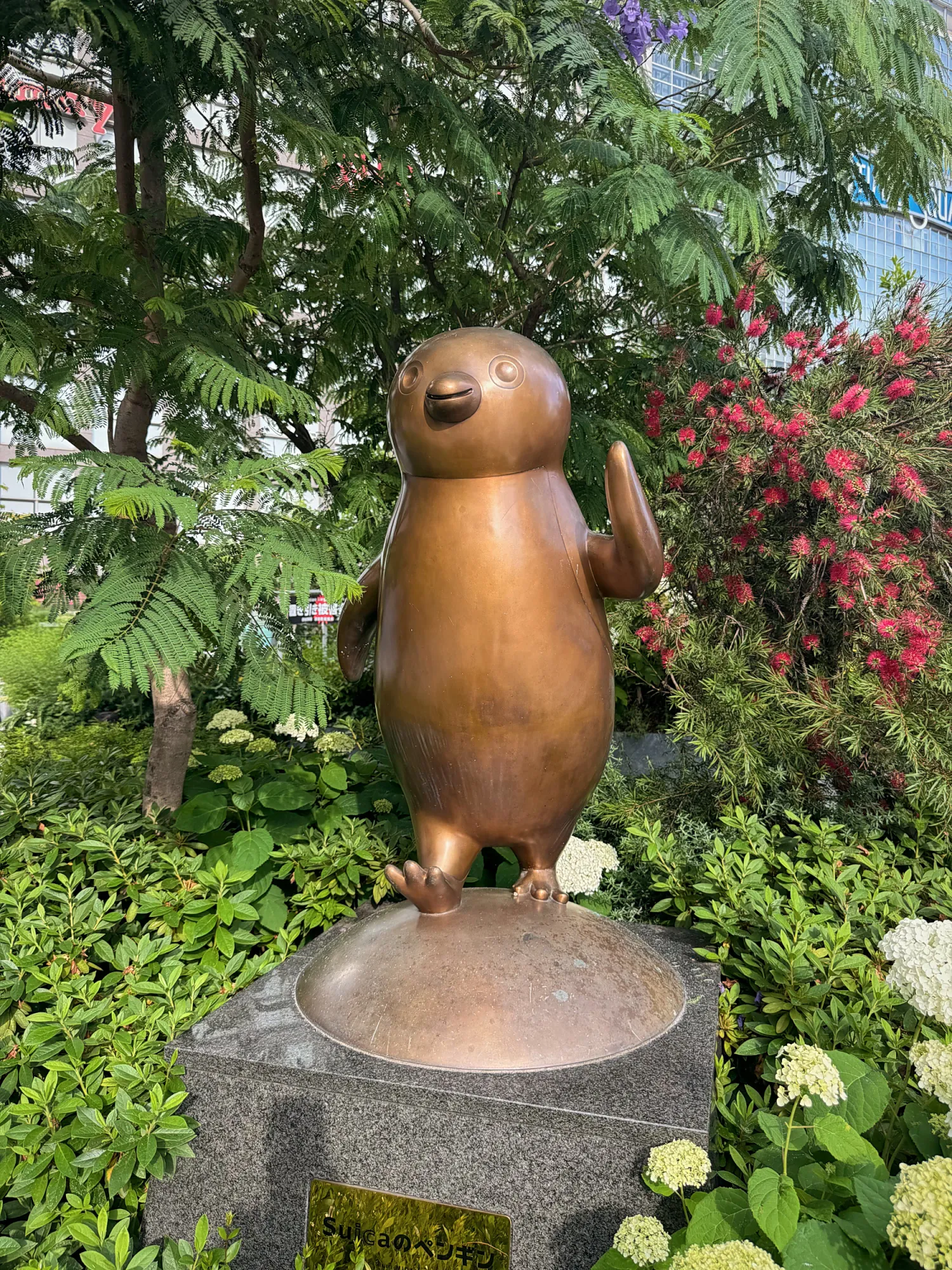 A bronze statue of the Suica penguin mascot standing on a stone base among green plants and pink flowers in Tokyo.