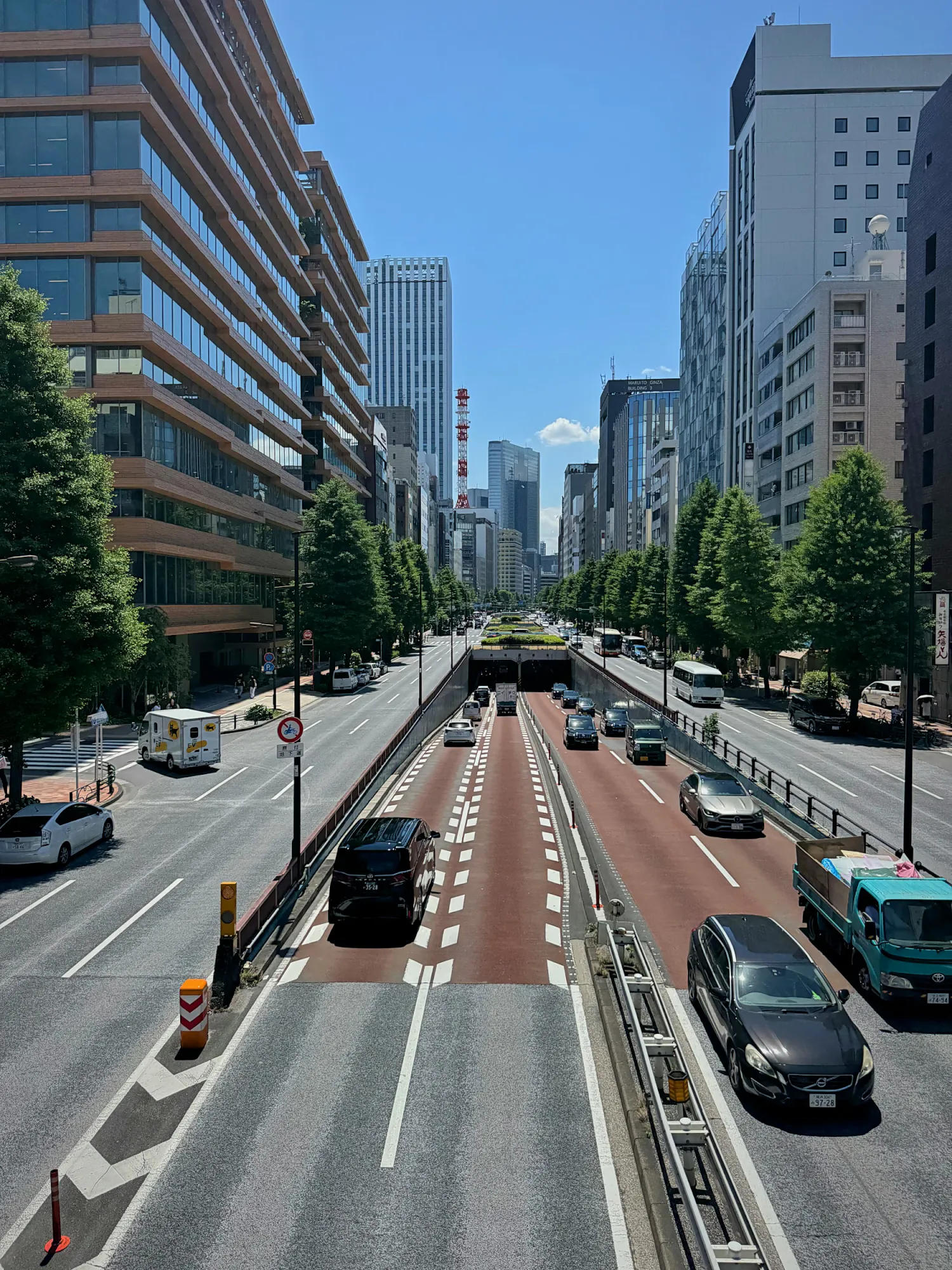 A wide Tokyo avenue lined with trees and office buildings under a clear blue sky.