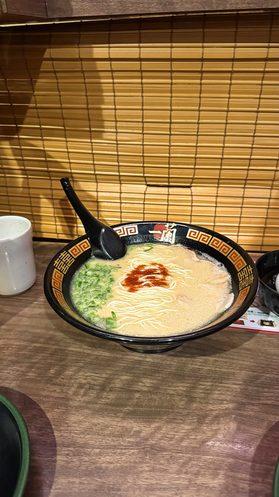 A bowl of Ichiran tonkotsu ramen with creamy broth, green onion, and a spoonful of red sauce on a wooden counter in a solo booth.
