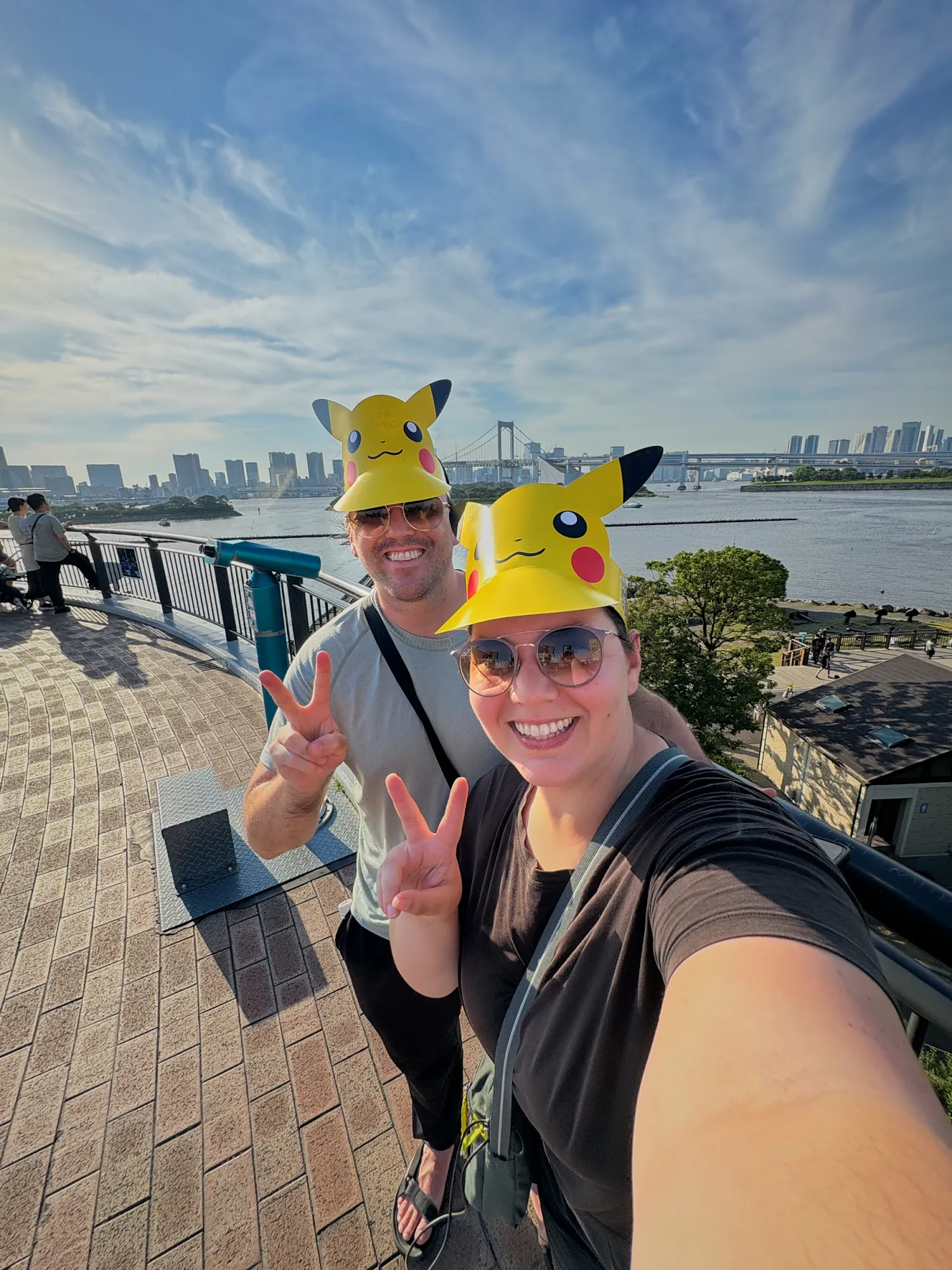 Bernie and Cream taking a selfie in yellow Pikachu visors on the waterfront promenade at Odaiba during Pokémon GO Fest Tokyo, with Tokyo Bay behind them.