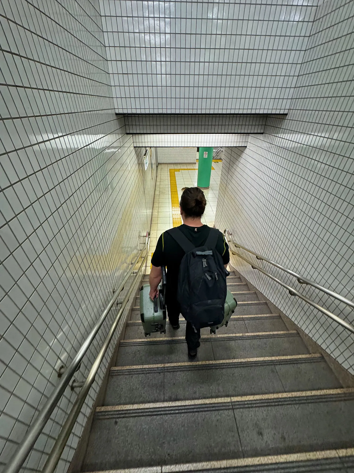 A traveler with a backpack carrying a suitcase down a long tiled staircase in a Tokyo subway station.