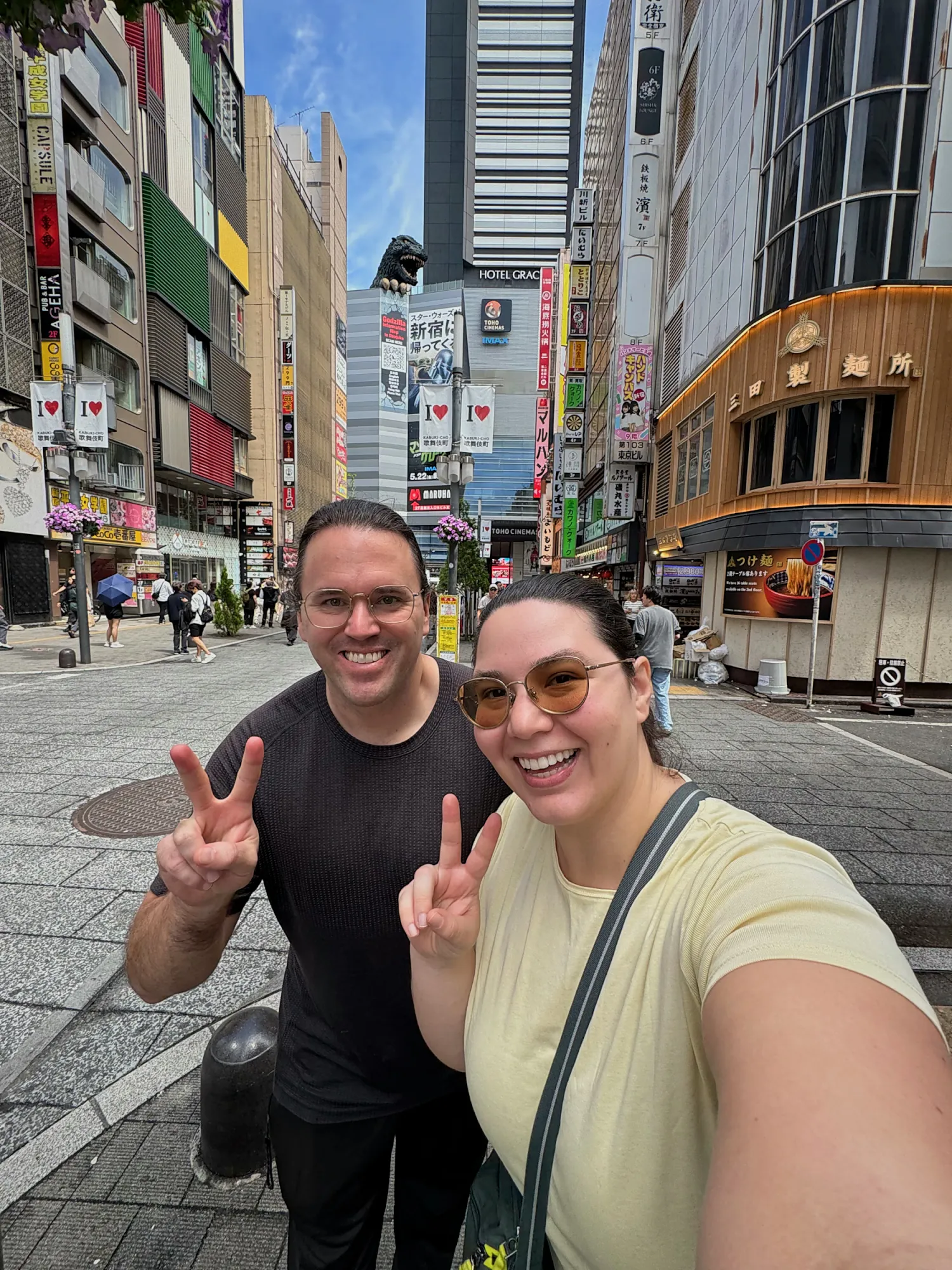 Bernie and Cream taking a selfie on Godzilla Road in Shinjuku, Tokyo, with the Godzilla head on top of a building behind them.