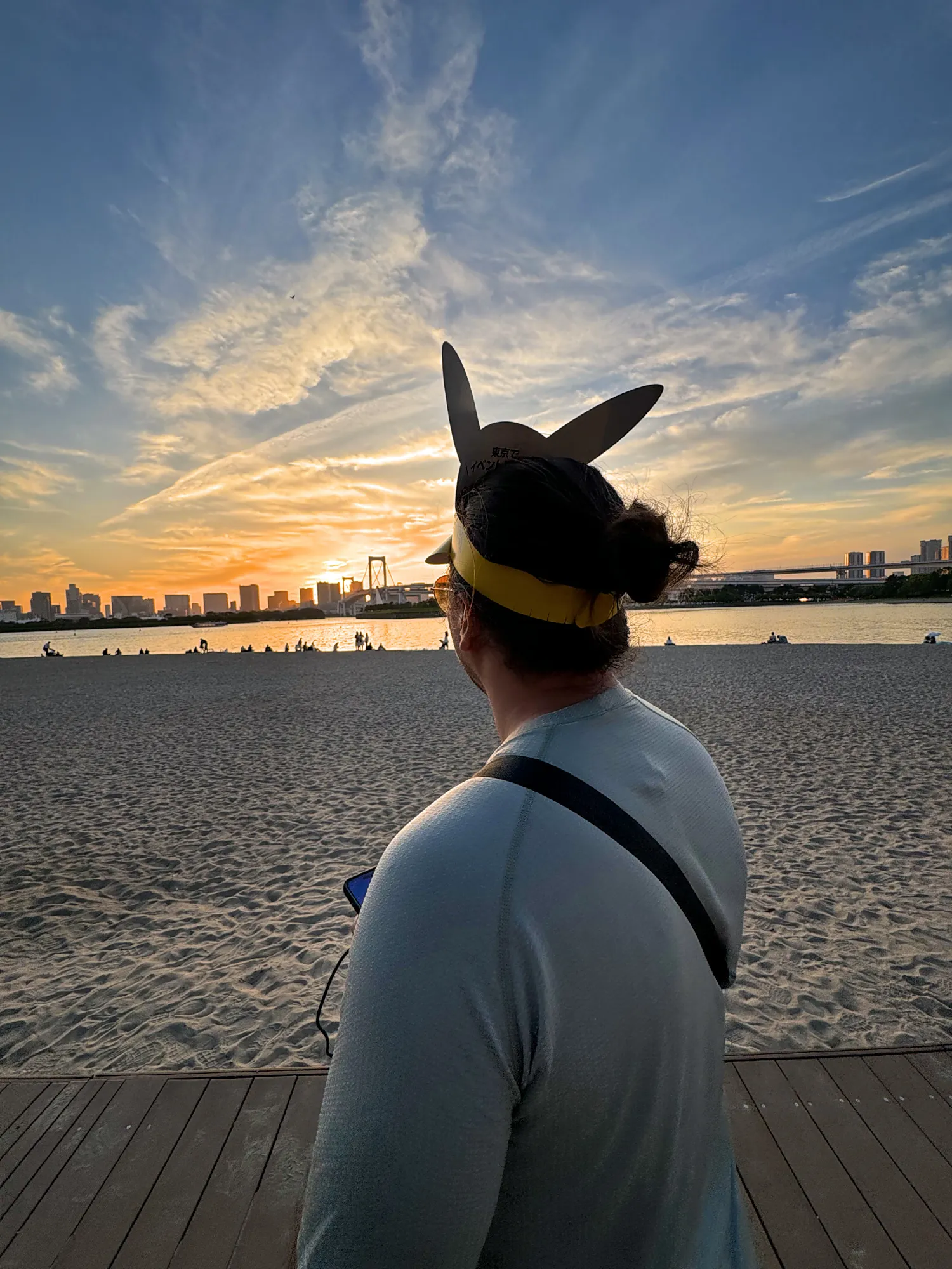 Bernie in a Pikachu visor watching the sunset over Tokyo Bay from the sand at Odaiba during Pokémon GO Fest Tokyo.