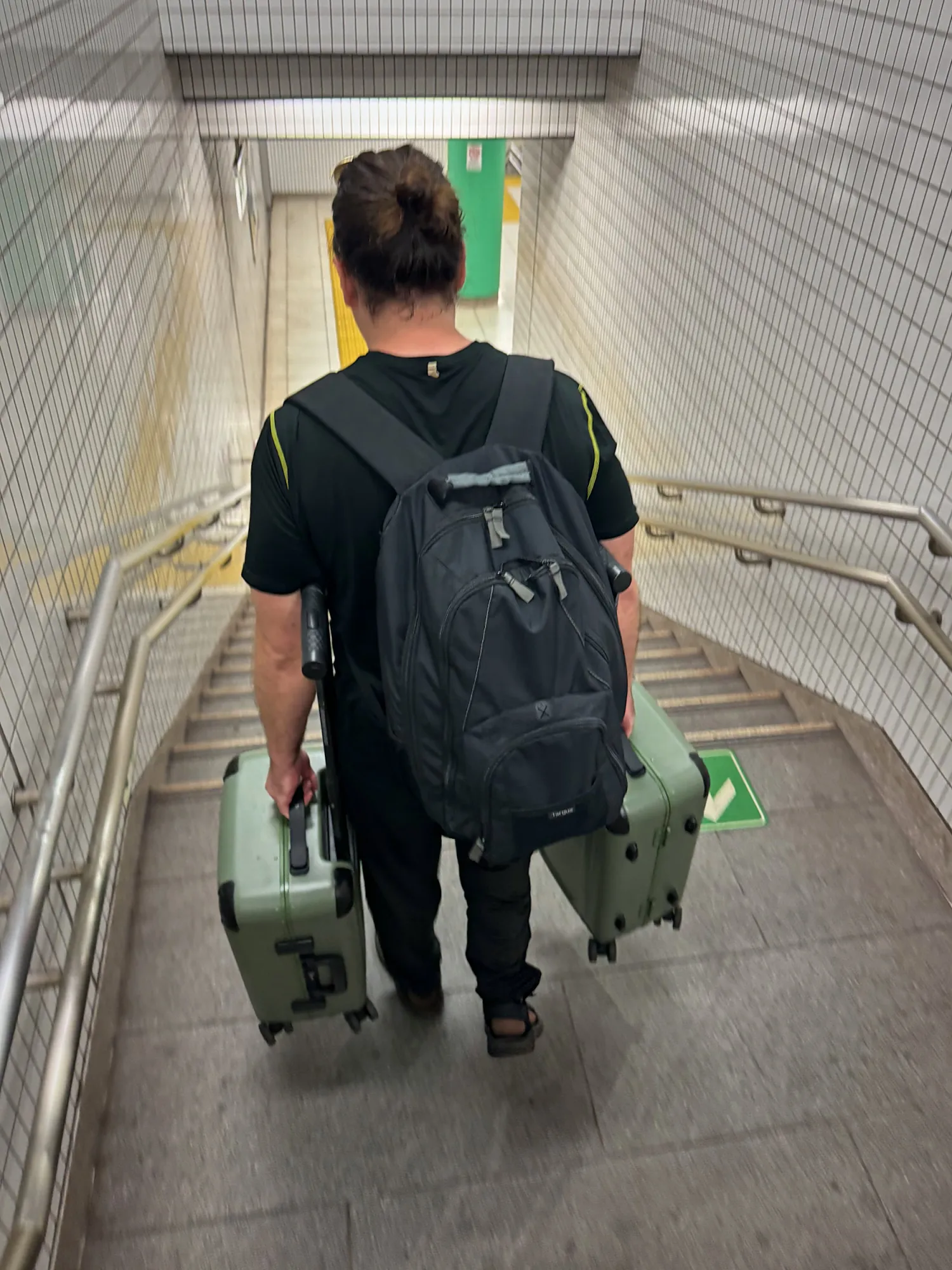 A traveler with a backpack carrying two green suitcases down a flight of stairs in a Tokyo subway station.