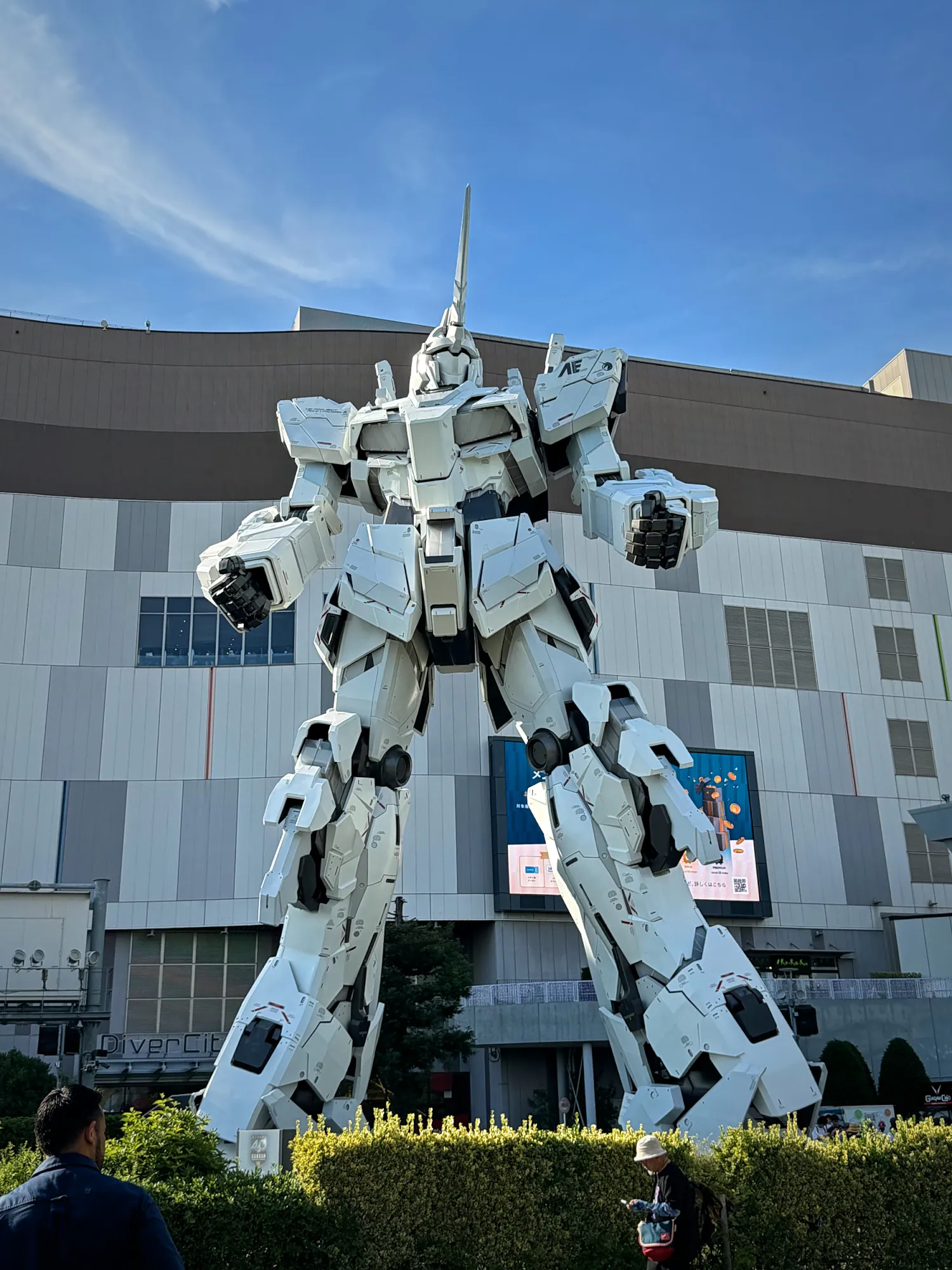 The life-size white Unicorn Gundam robot statue standing outside a shopping mall in Odaiba, Tokyo, under a blue sky, in May 2026.