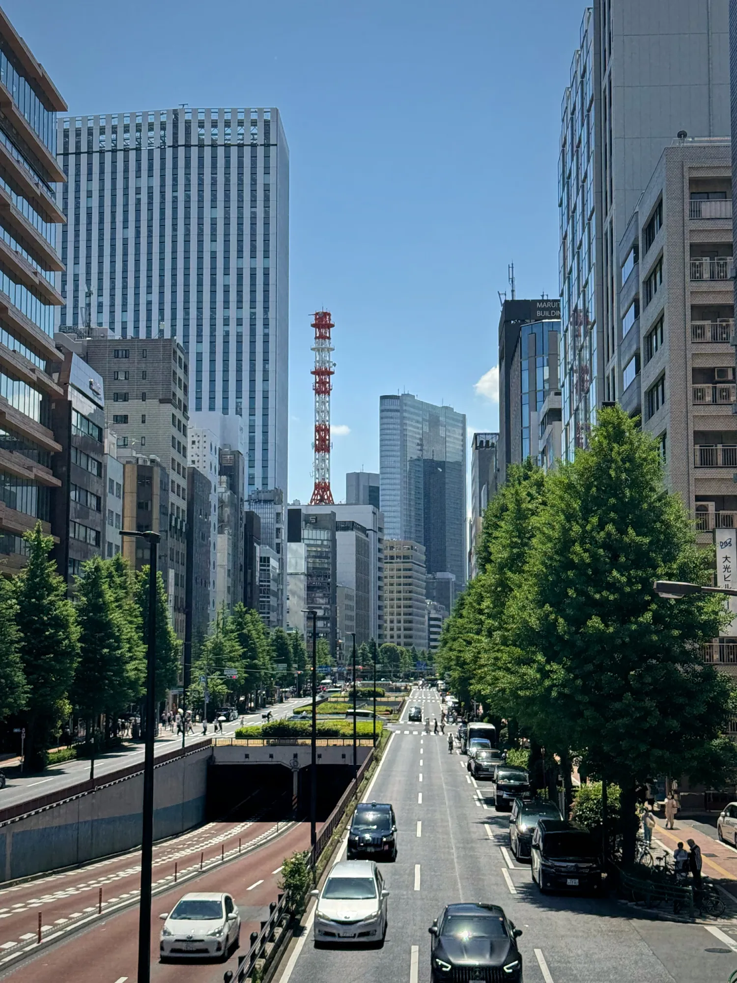 A wide, tree-lined Tokyo avenue running between office towers on a sunny day, with the red and white Tokyo Tower in the distance.