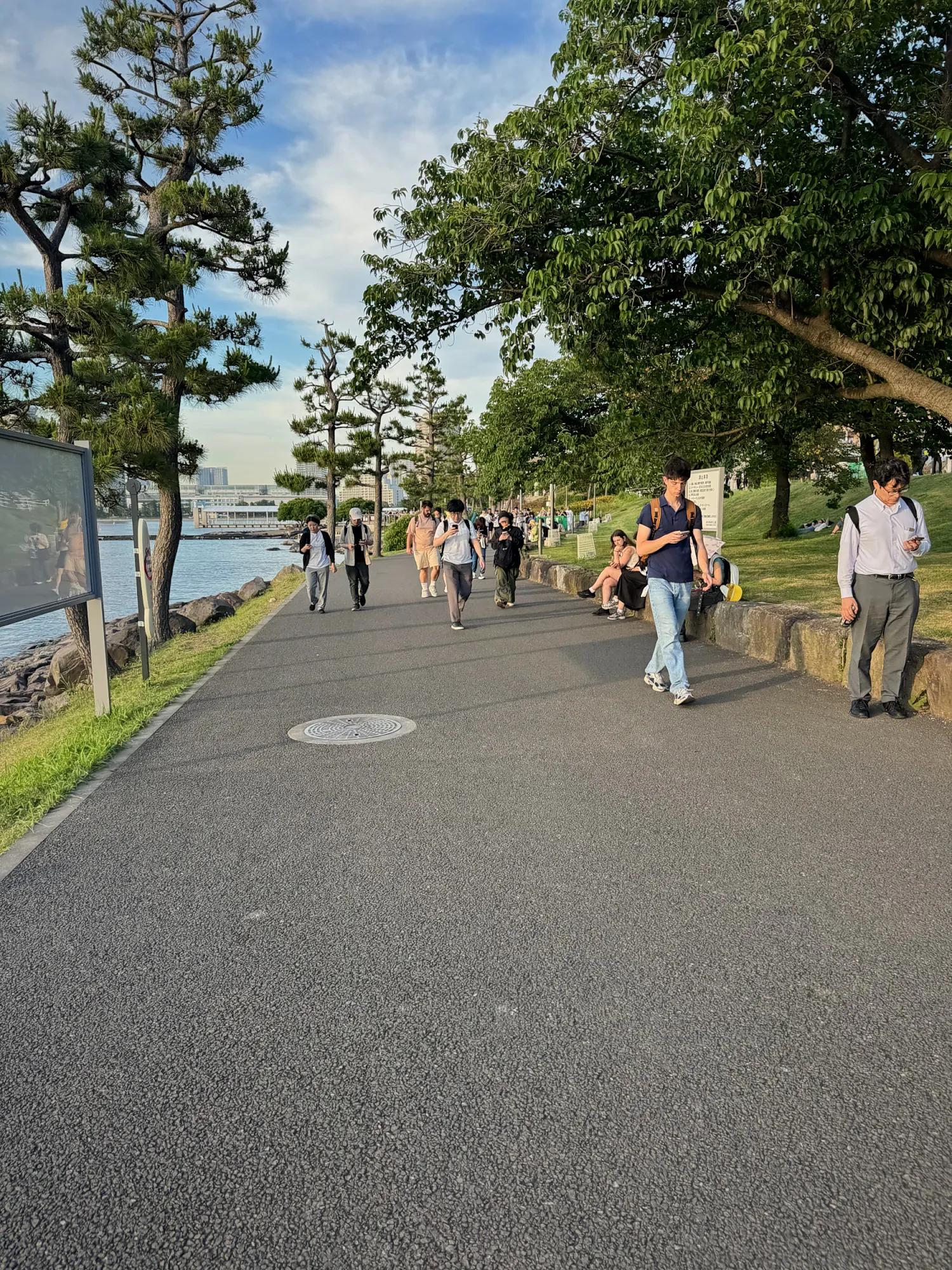 People walking along a tree-lined waterfront promenade in Odaiba, Tokyo, in the late afternoon.