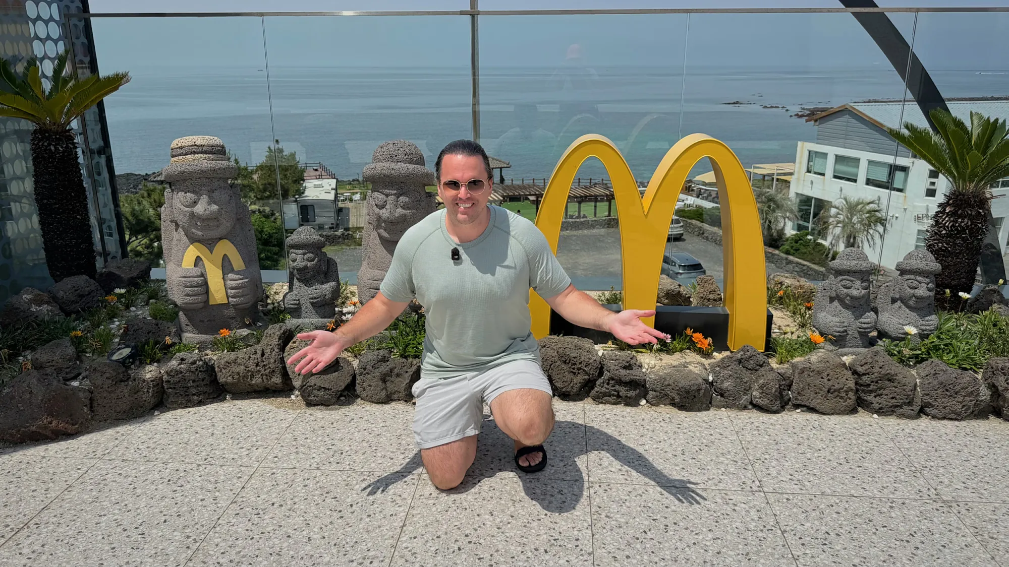 A traveler kneeling with arms open by the golden arches on the ocean-view rooftop of a McDonald's on Jeju Island, South Korea.