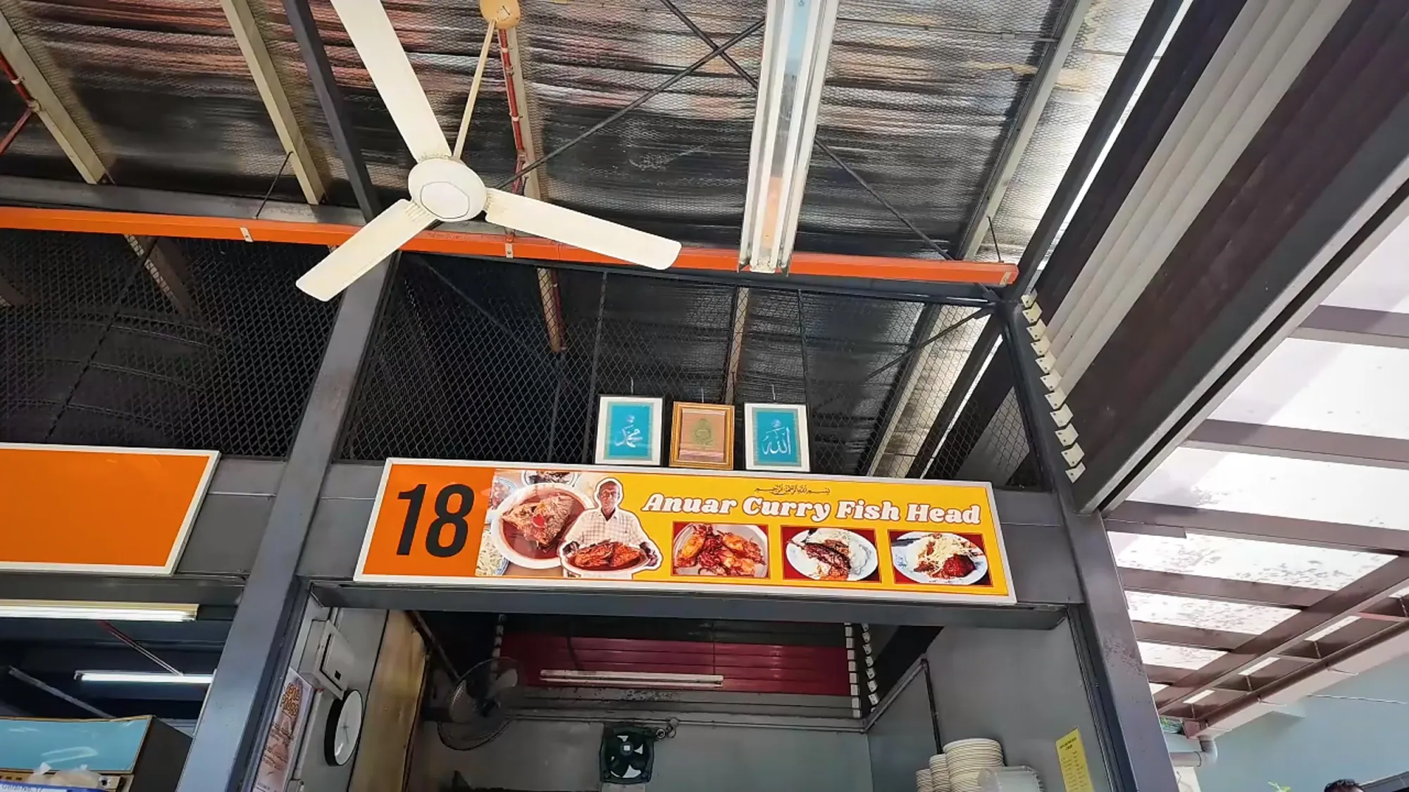The orange-and-yellow stall 18 sign reading Anuar Curry Fish Head under a metal roof with a ceiling fan in Bangsar, Kuala Lumpur.