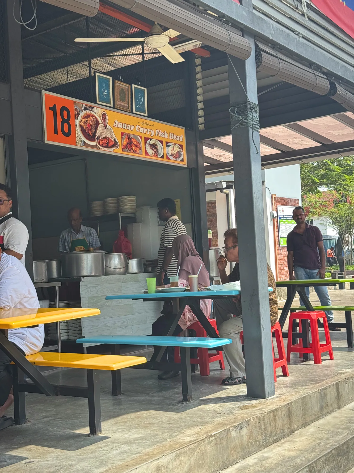 The Anuar Curry Fish Head stall with yellow and blue tables in an open-air food court in Lucky Garden, Bangsar, Kuala Lumpur.