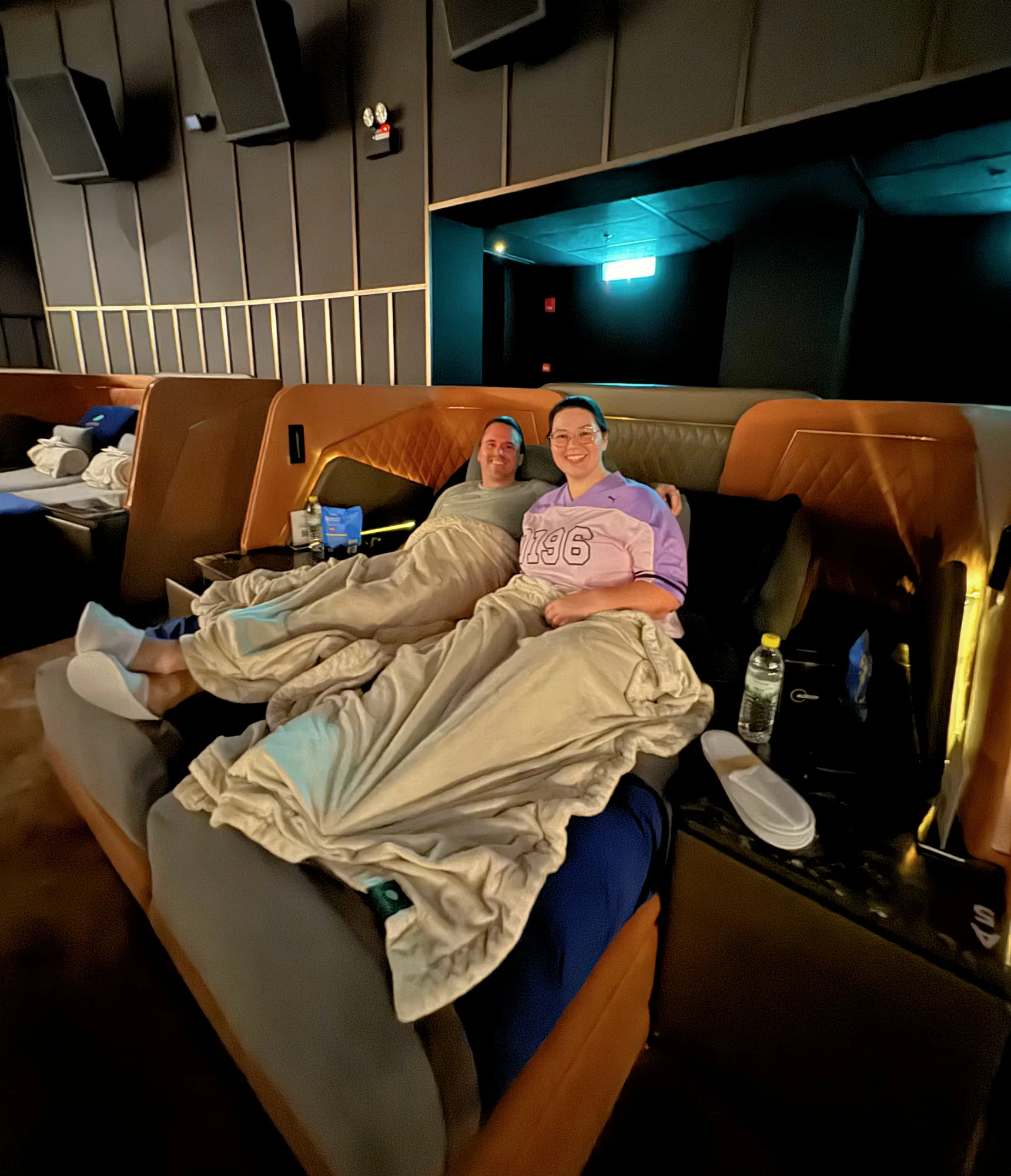 A smiling couple relaxes under blankets in plush recliner beds inside a Getha Lux Suite at Aurum Theatre, The Exchange TRX, in Kuala Lumpur.