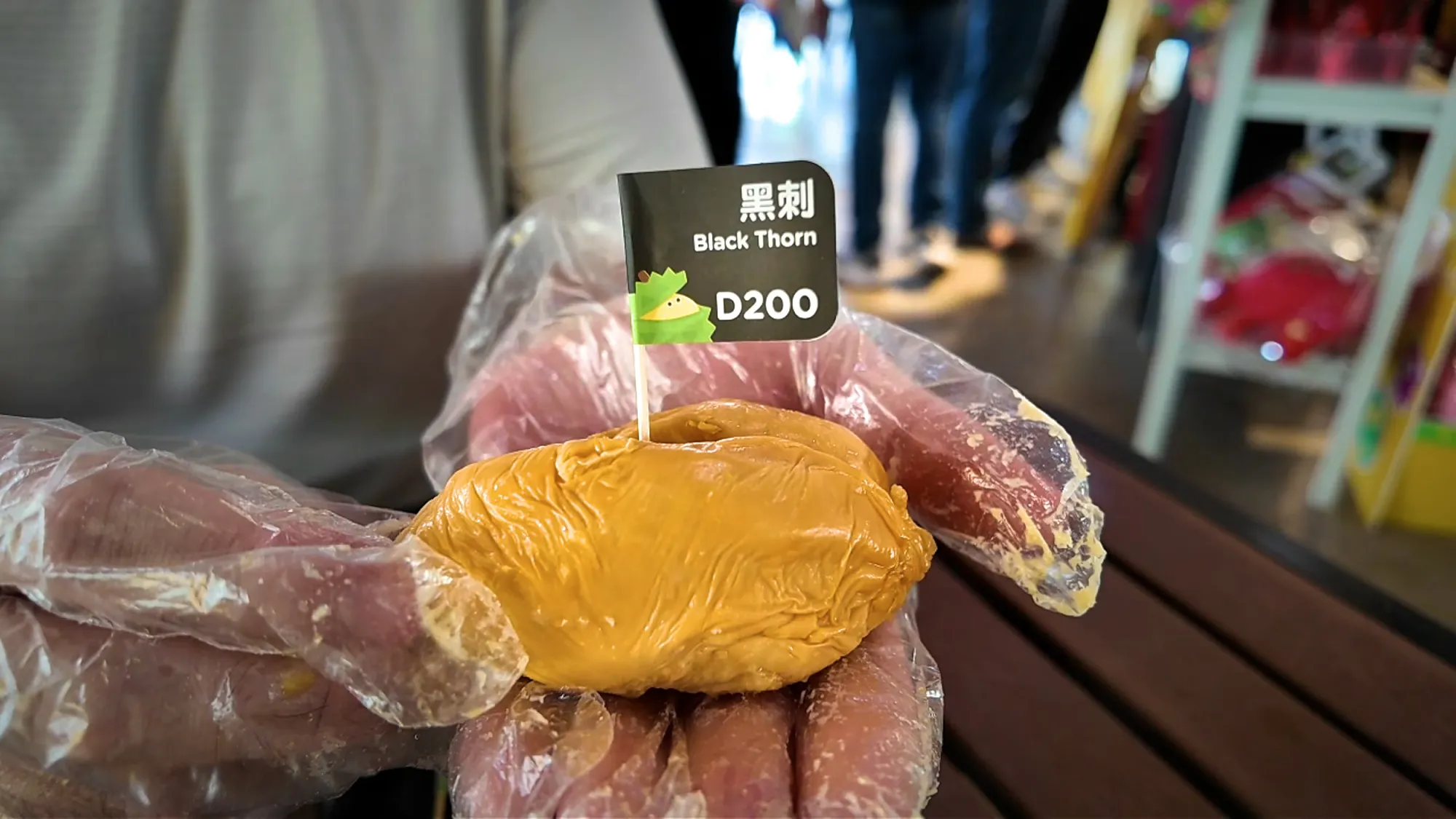 A gloved hand holding a deep orange Black Thorn durian piece with a black flag marked D200.