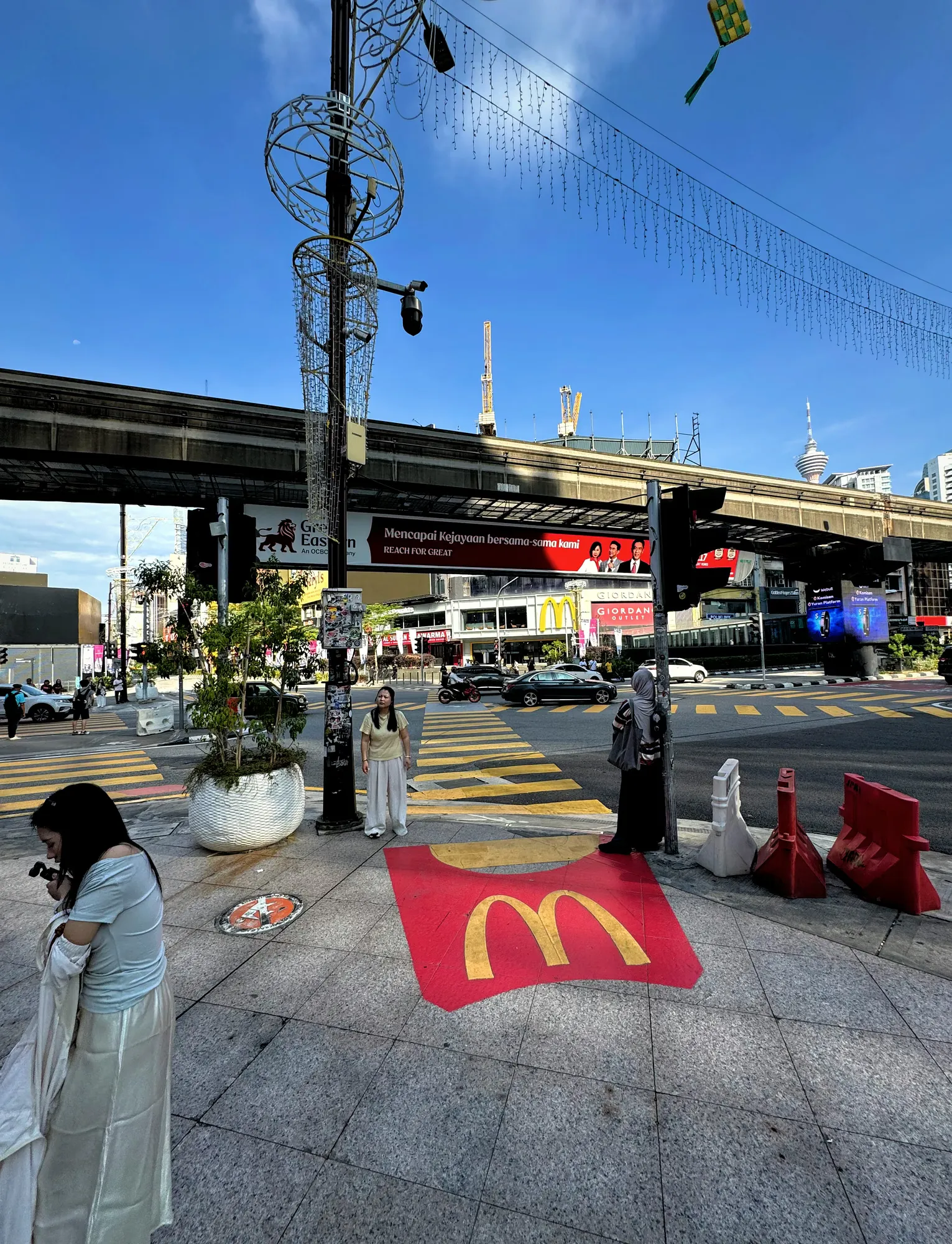 A wide angle view of the bustling Bukit Bintang intersection under a blue sky, showing traffic, monorail tracks, and a painted McDonald's crosswalk graphic.