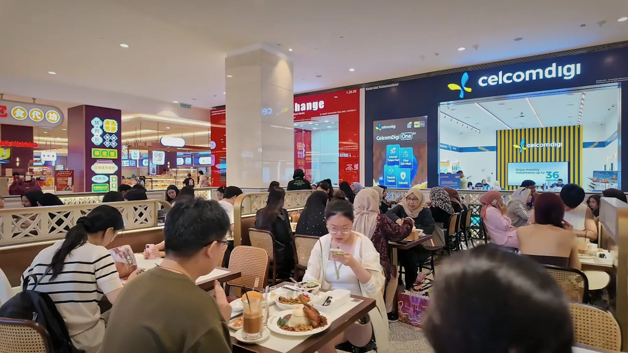 Diners at tables in a brightly lit shopping mall food court in Kuala Lumpur.