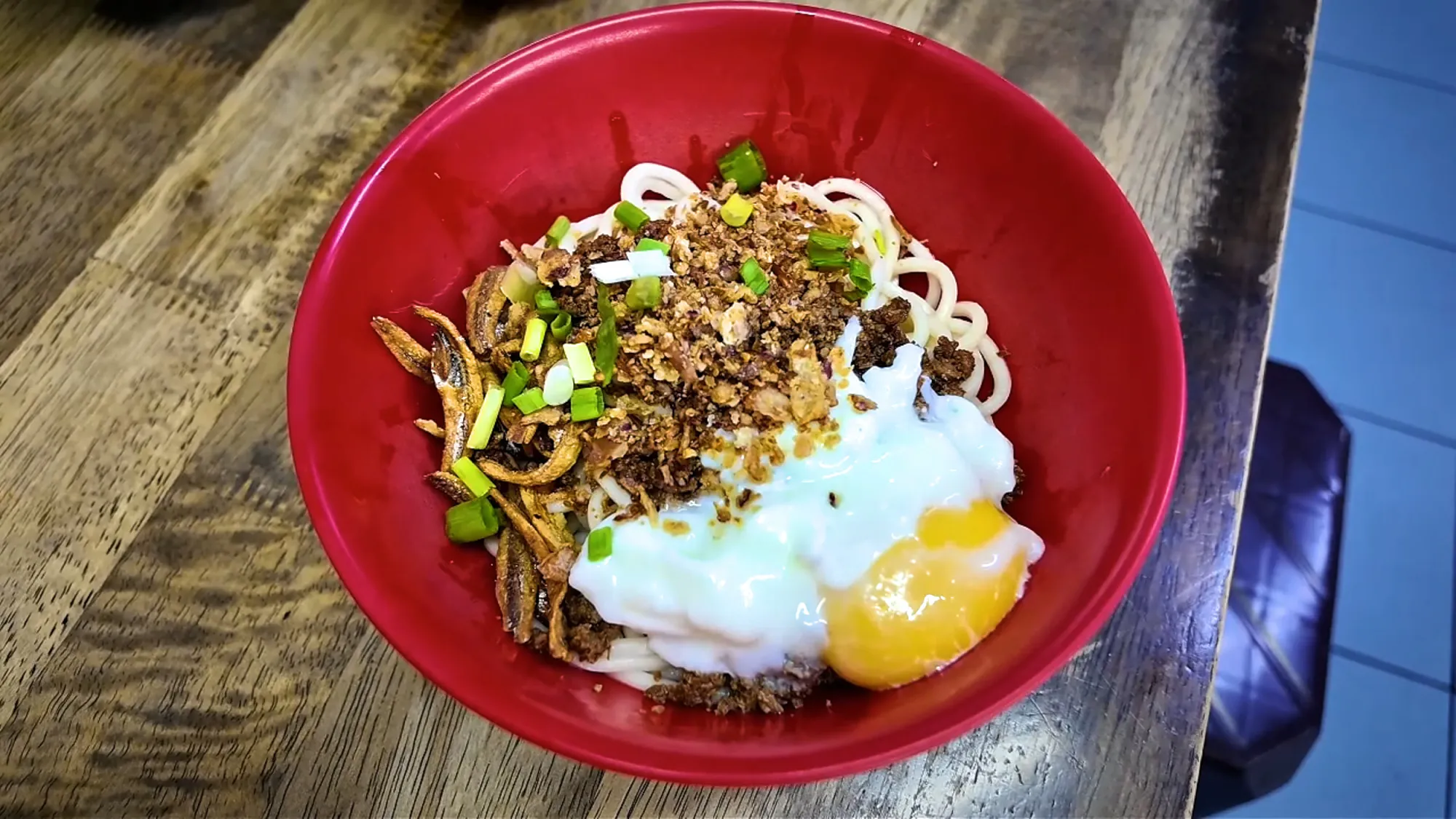A red bowl of dry chili pan mee topped with a poached egg, minced pork, and crispy anchovies at Kin Kin