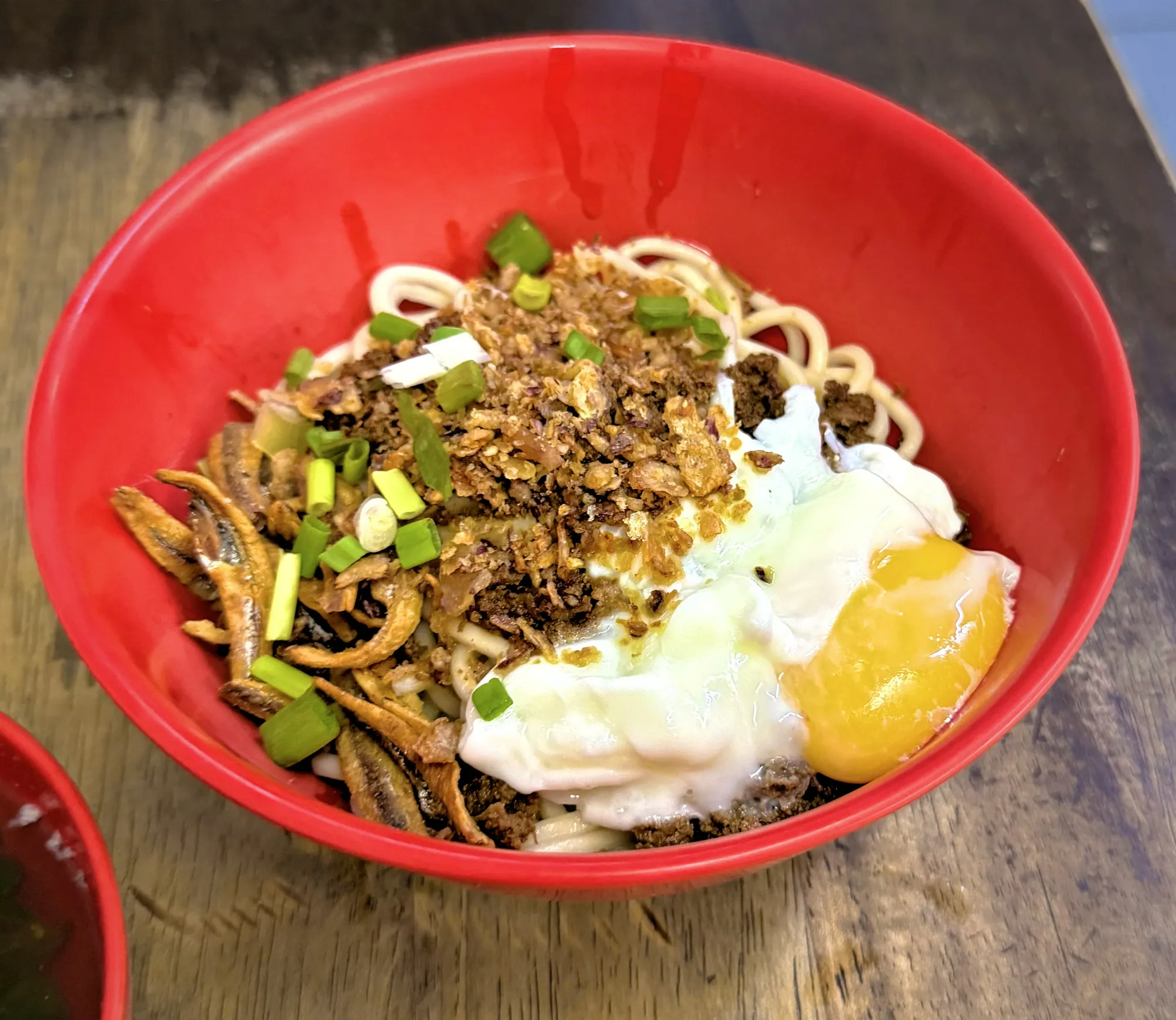 A red bowl of dry chili pan mee topped with a poached egg, minced pork, crispy anchovies, and spring onions at Restoran Kin Kin in Kuala Lumpur.