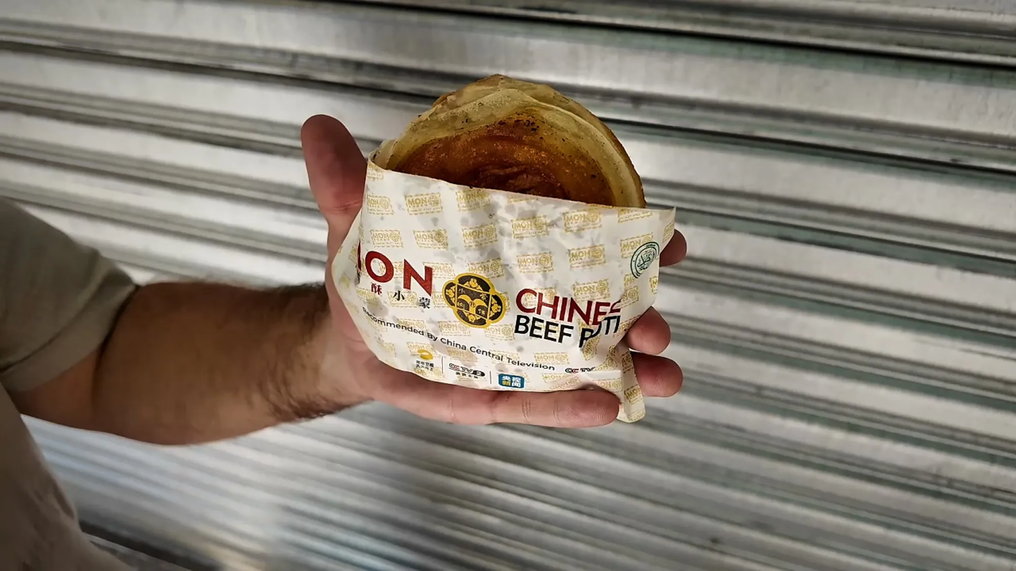 Hands holding a round, golden, crispy Mon Chinese Beef Roti in its paper wrapper in Kuala Lumpur.