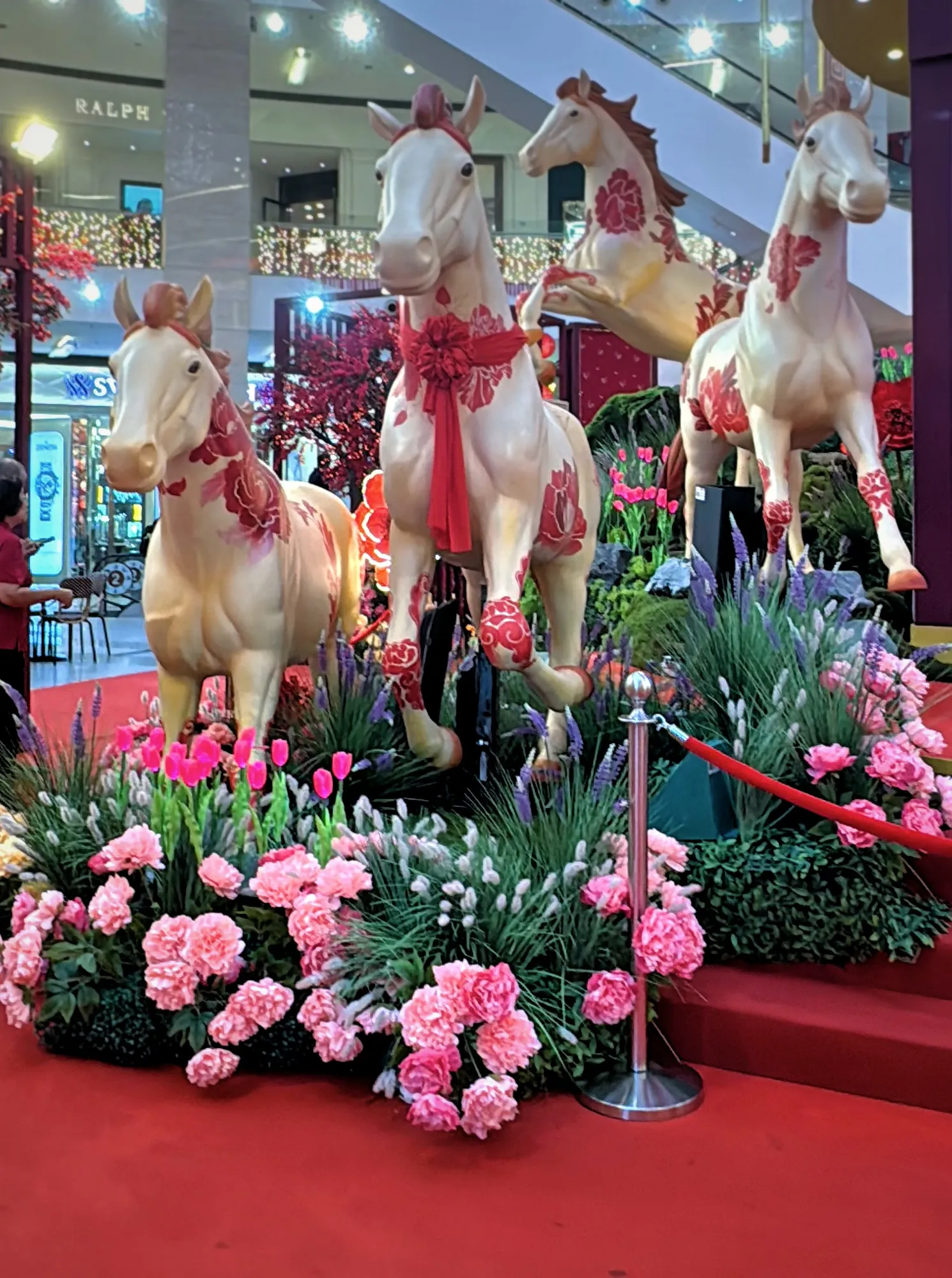 White horse sculptures with crimson floral patterns among pink flowers, a Year of the Horse display inside Pavilion Kuala Lumpur.
