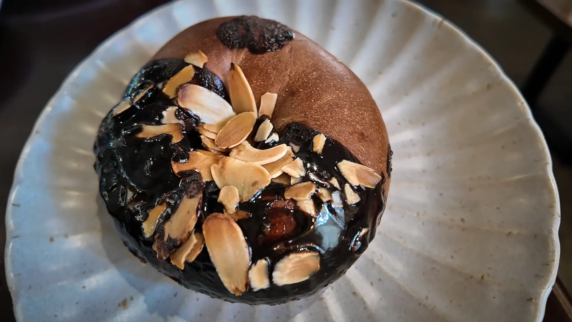 Chocolate bagel topped with a glossy chocolate glaze and toasted almond slices on a white plate.