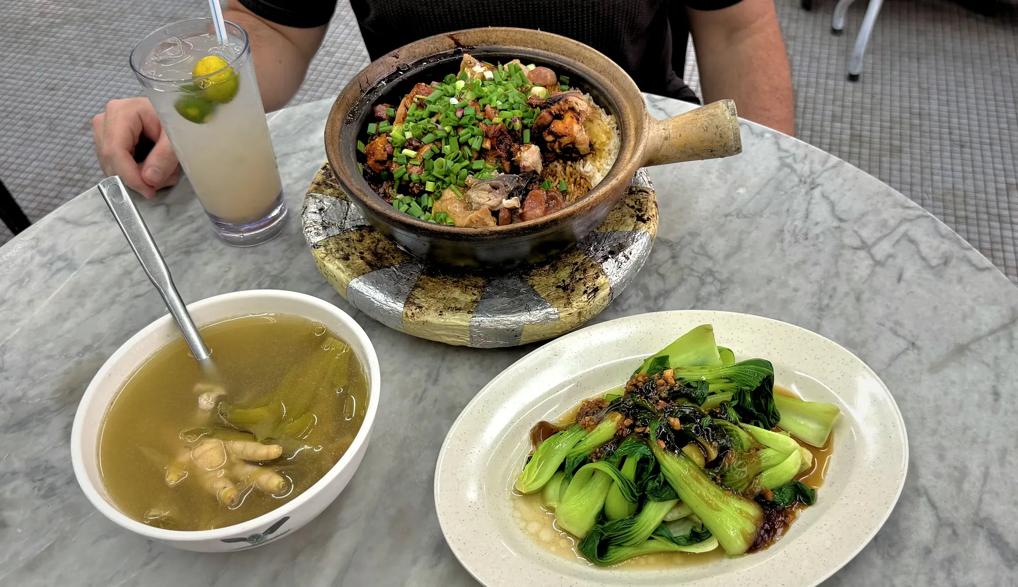 Claypot chicken rice topped with spring onions, served with bok choy, herbal chicken feet soup, and a lime juice on a marble table at 168 Claypot Chicken Rice in Kuala Lumpur