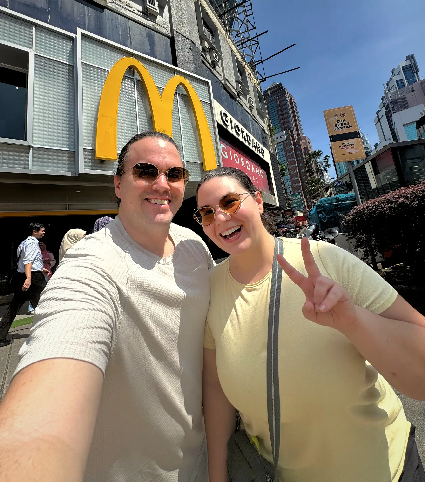 A smiling couple takes a sunny outdoor selfie in front of the iconic giant McDonald's golden arches in Bukit Bintang, Kuala Lumpur.