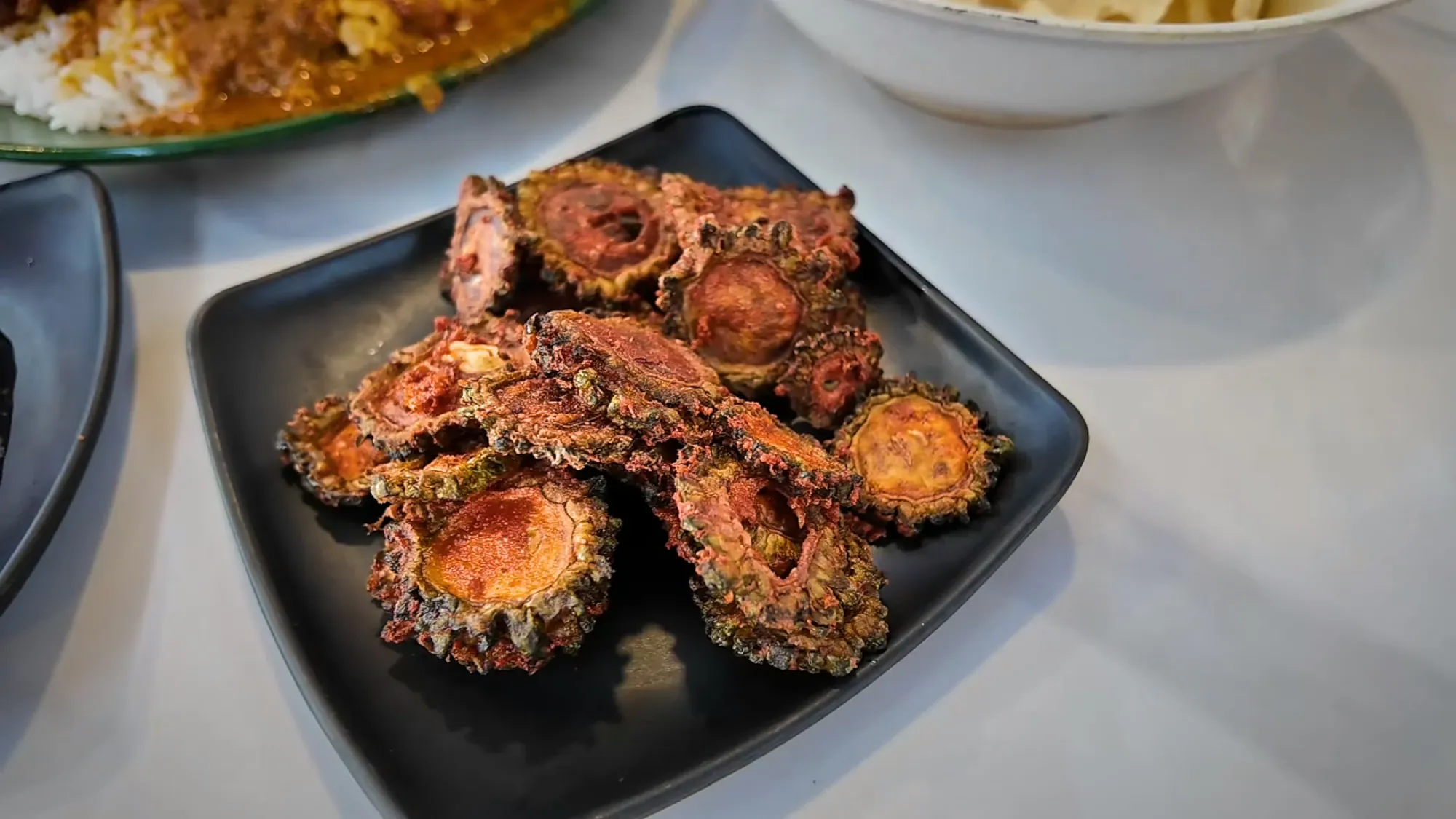 Crispy fried bitter gourd slices on a small black plate at Line Clear in Kampung Baru, Kuala Lumpur.