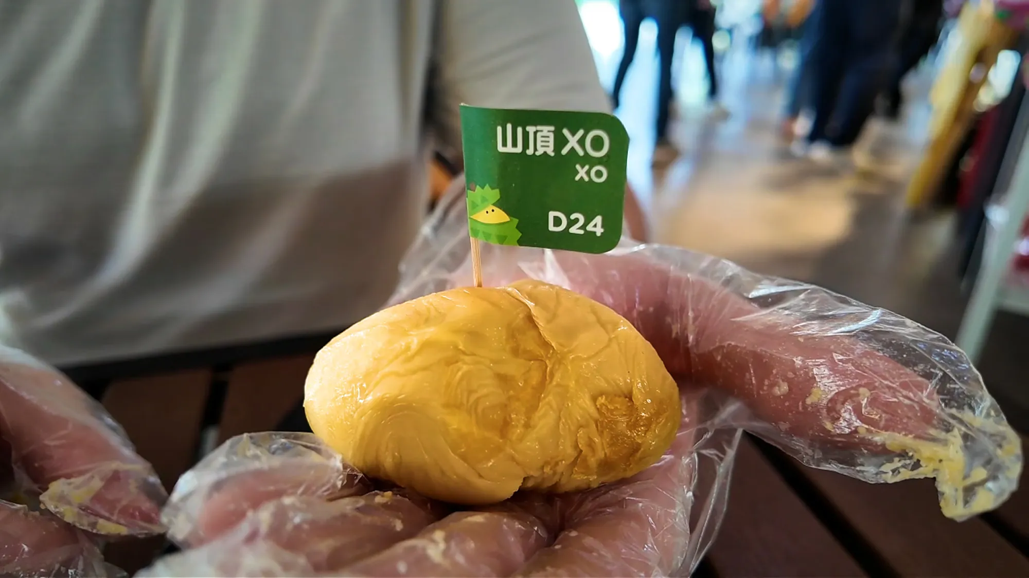 A gloved hand holding a round, pale golden XO durian piece with a green flag marked XO and D24.