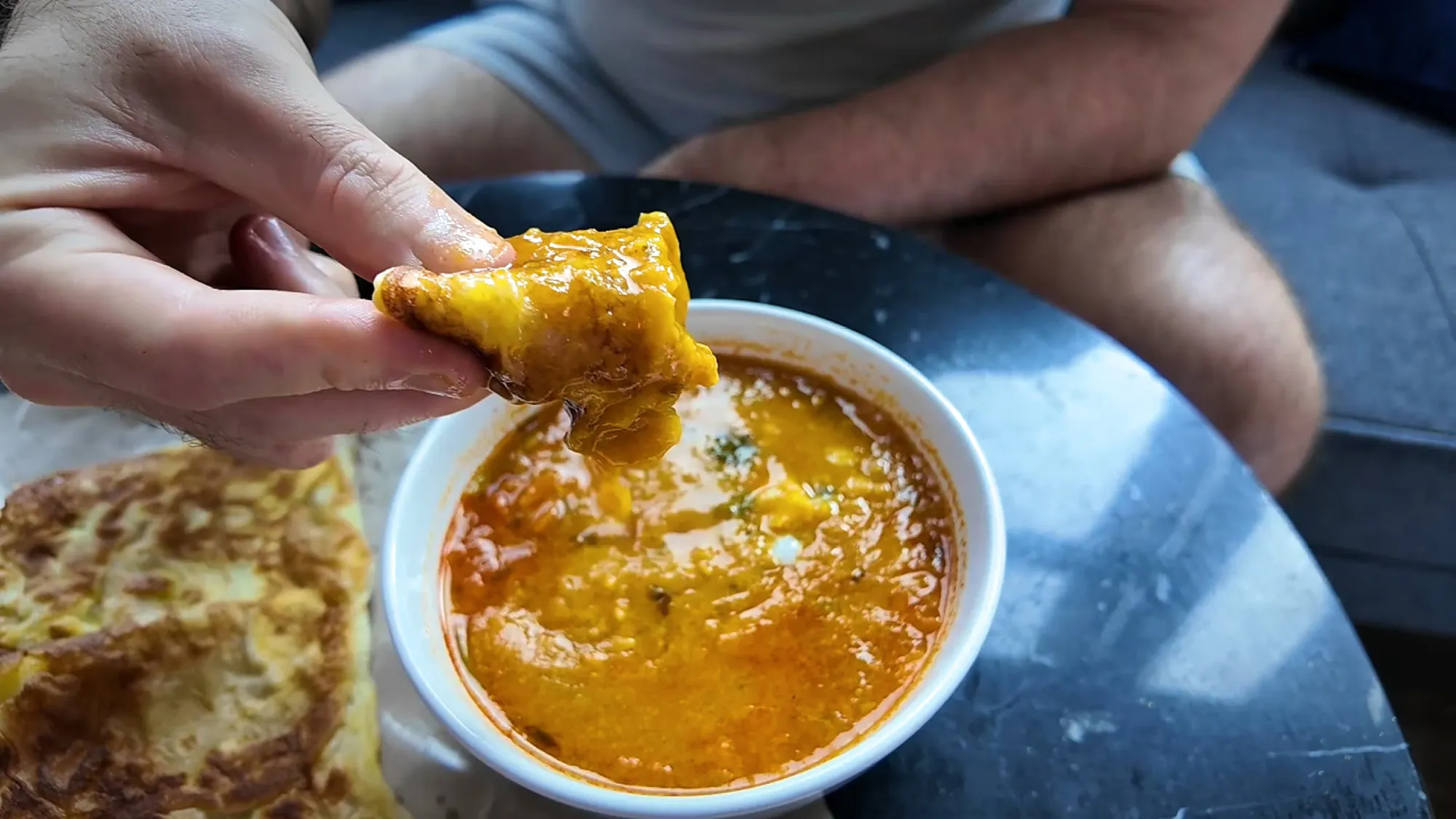 A piece of roti being dunked into a bowl of thick orange lentil dhal, with a roti telur on the side