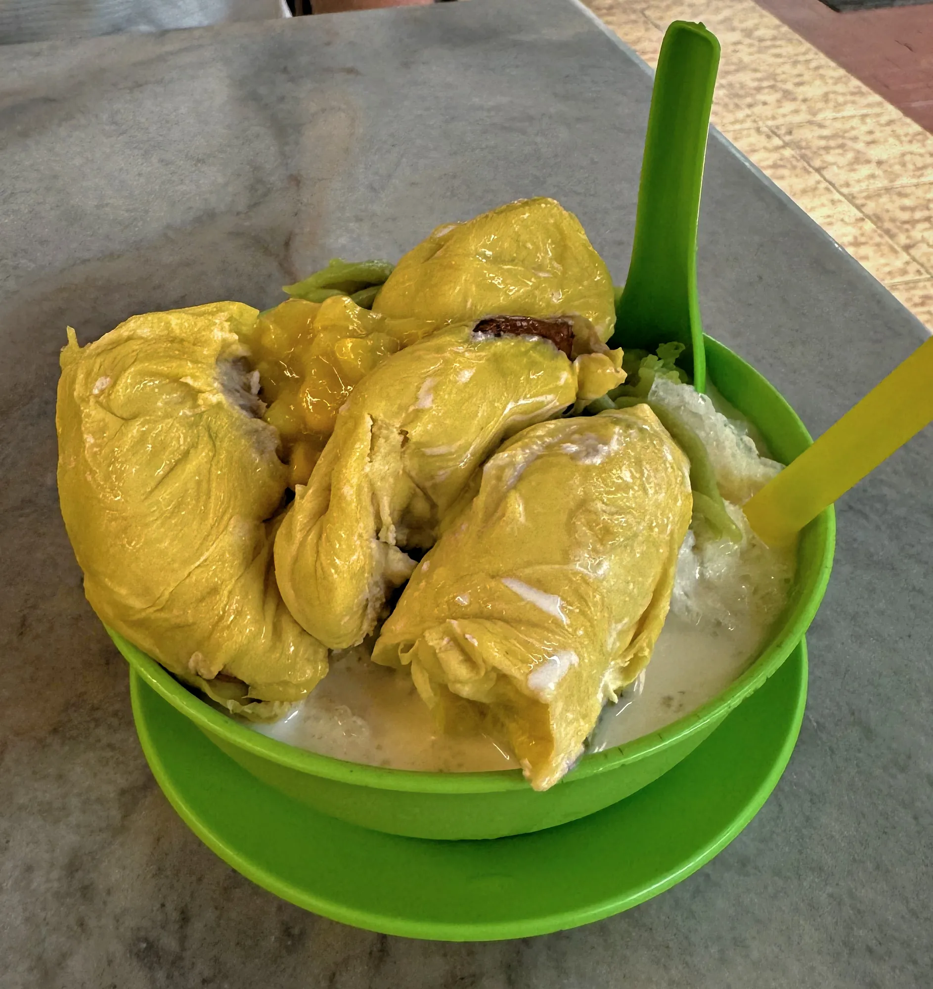 A green bowl piled with yellow durian pulp, shaved ice, coconut milk, and cendol jelly on a marble table in Kuala Lumpur