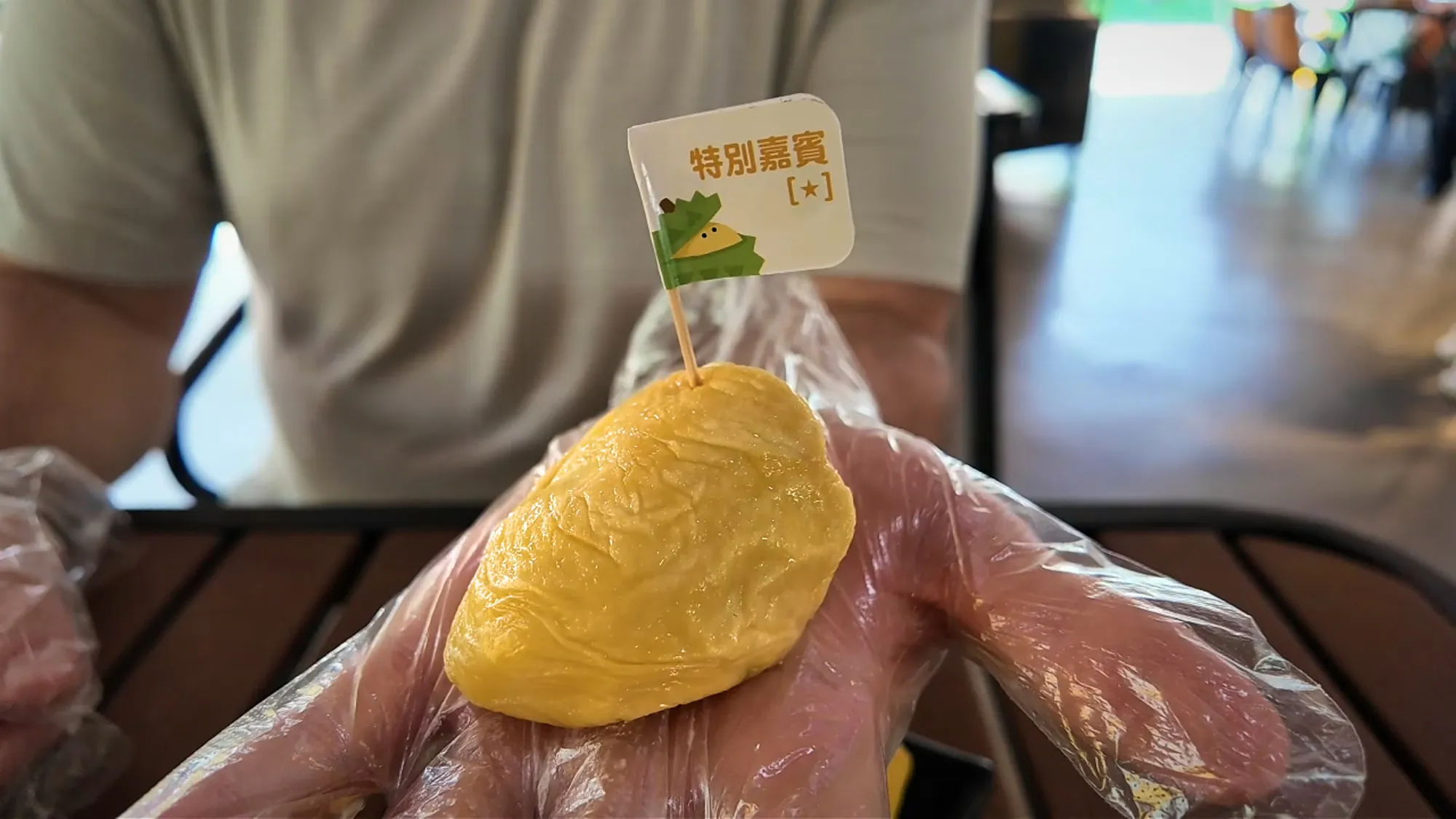 A gloved hand holding a pale yellow durian piece with a flag reading 特別嘉賓 and a star.
