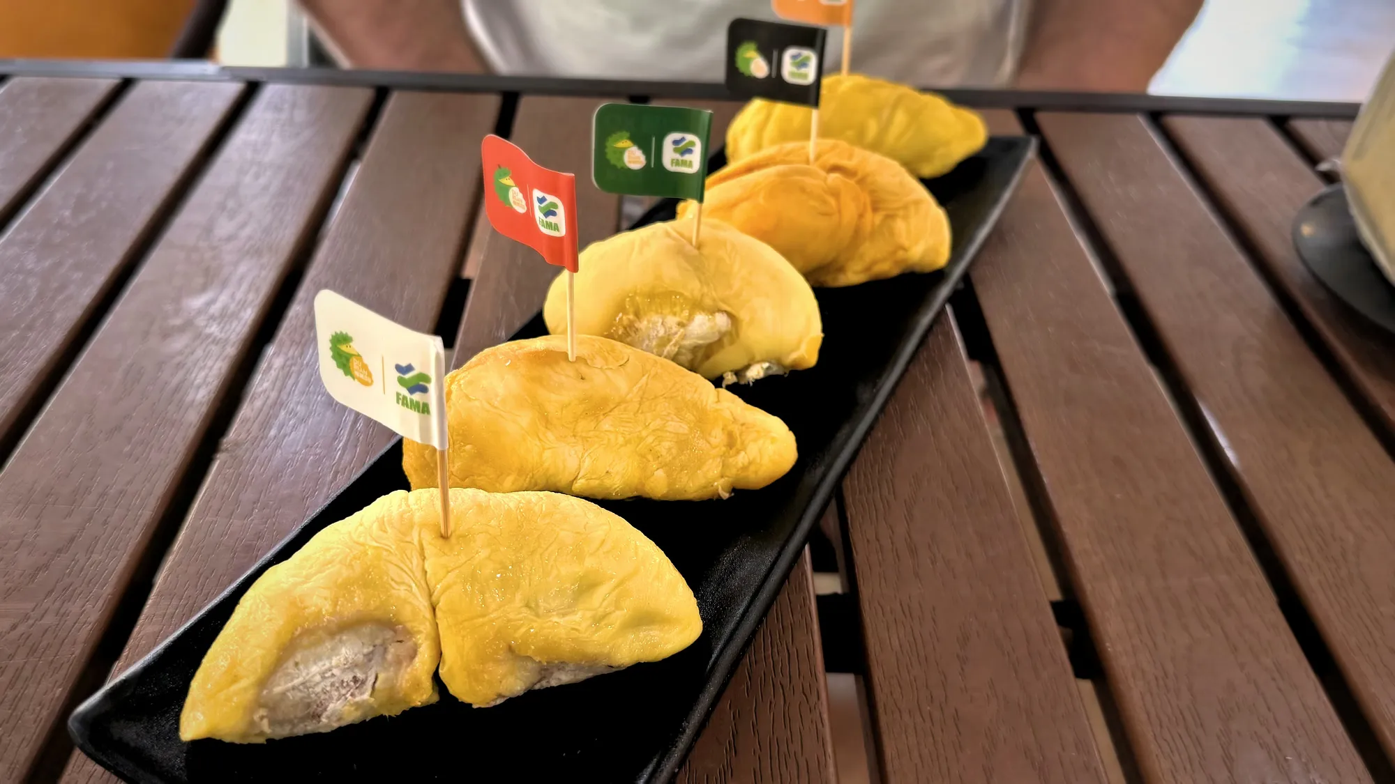 Five durian segments on a long black platter, each with a small colored flag, on a wooden table at DurianBB Park, Kuala Lumpur.