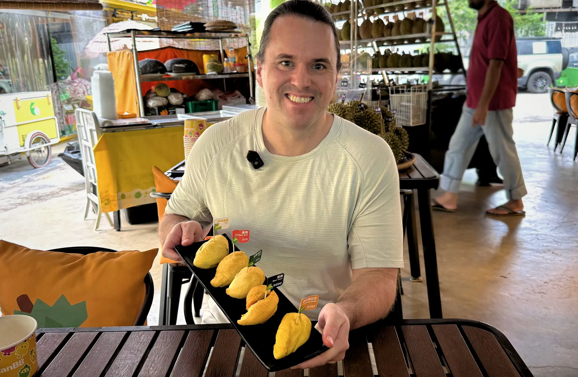 A smiling man presents a flight of five distinct Malaysian durian varieties on a black slate platter at DurianBB Park in Kuala Lumpur.