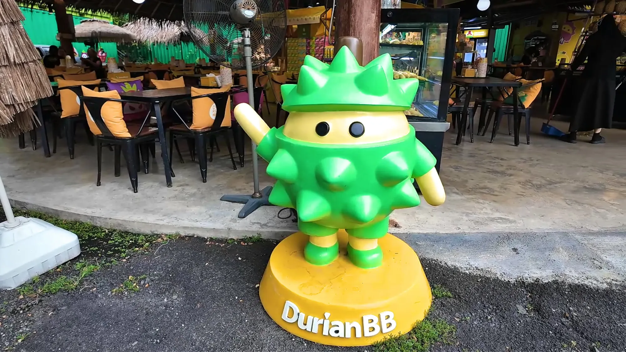 A bright green spiky durian mascot statue on a yellow DurianBB base in front of the seating area.