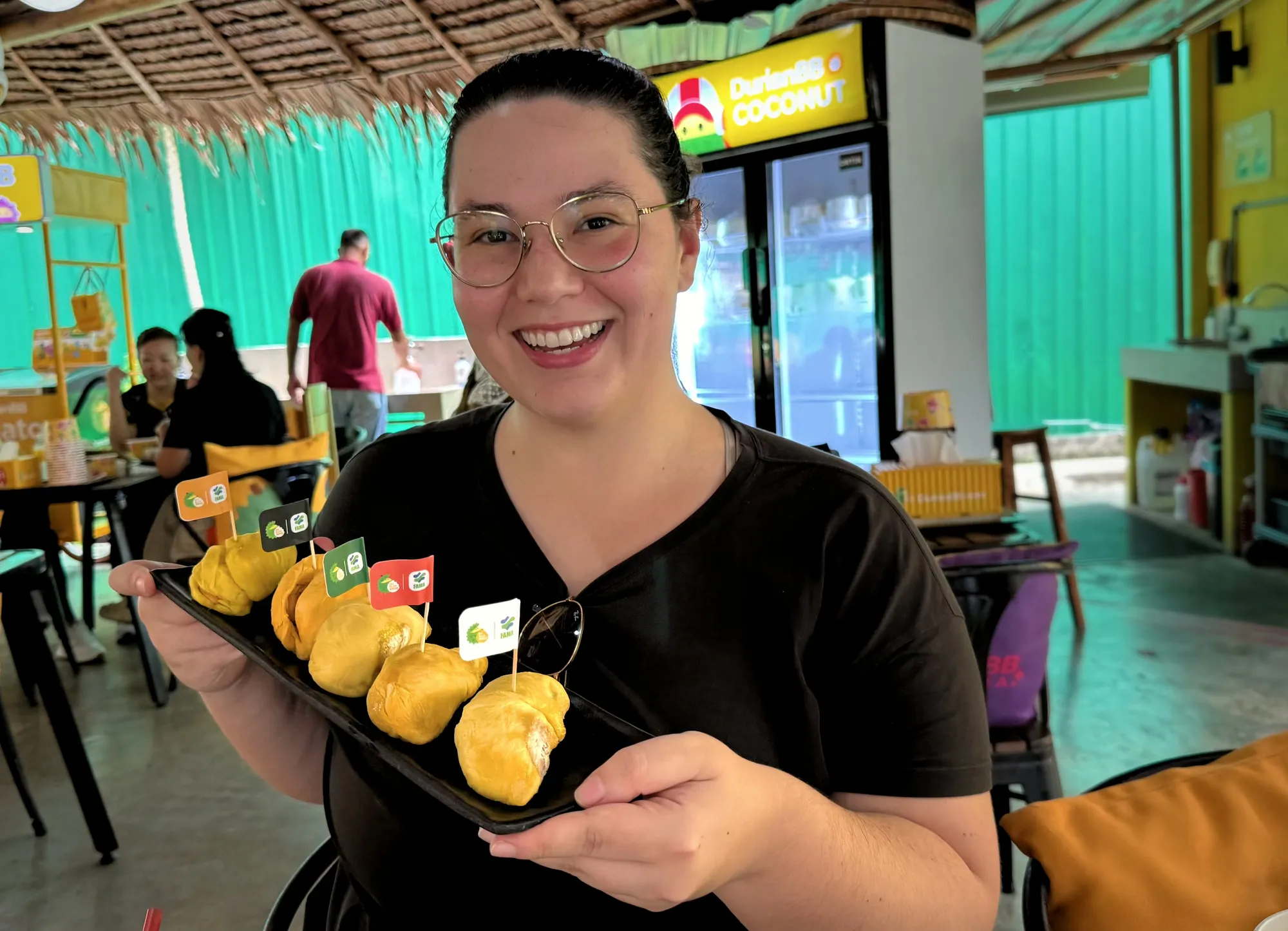 A smiling woman wearing glasses holds a long black plate featuring five golden Malaysian durian cultivars at DurianBB Park in Kuala Lumpur.