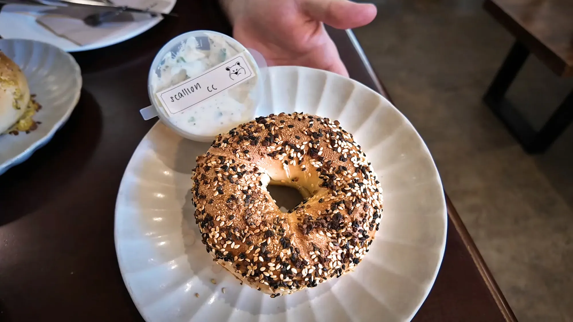 Everything bagel covered in sesame and poppy seeds on a white plate, with a tub of scallion cream cheese behind it.
