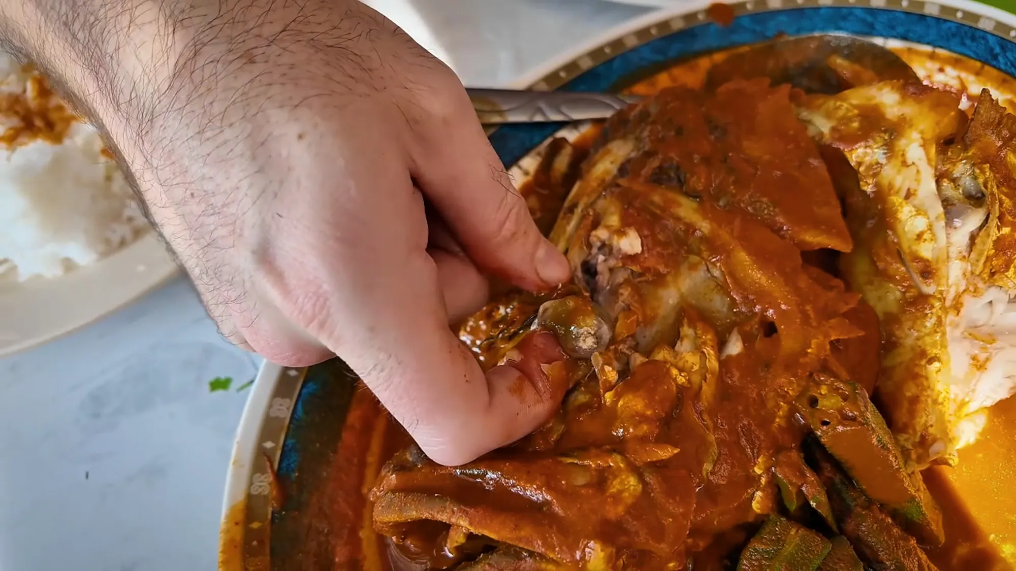 Close-up of a hand pulling meat from a fish head in thick red curry with okra on a blue-rimmed platter in Kuala Lumpur.