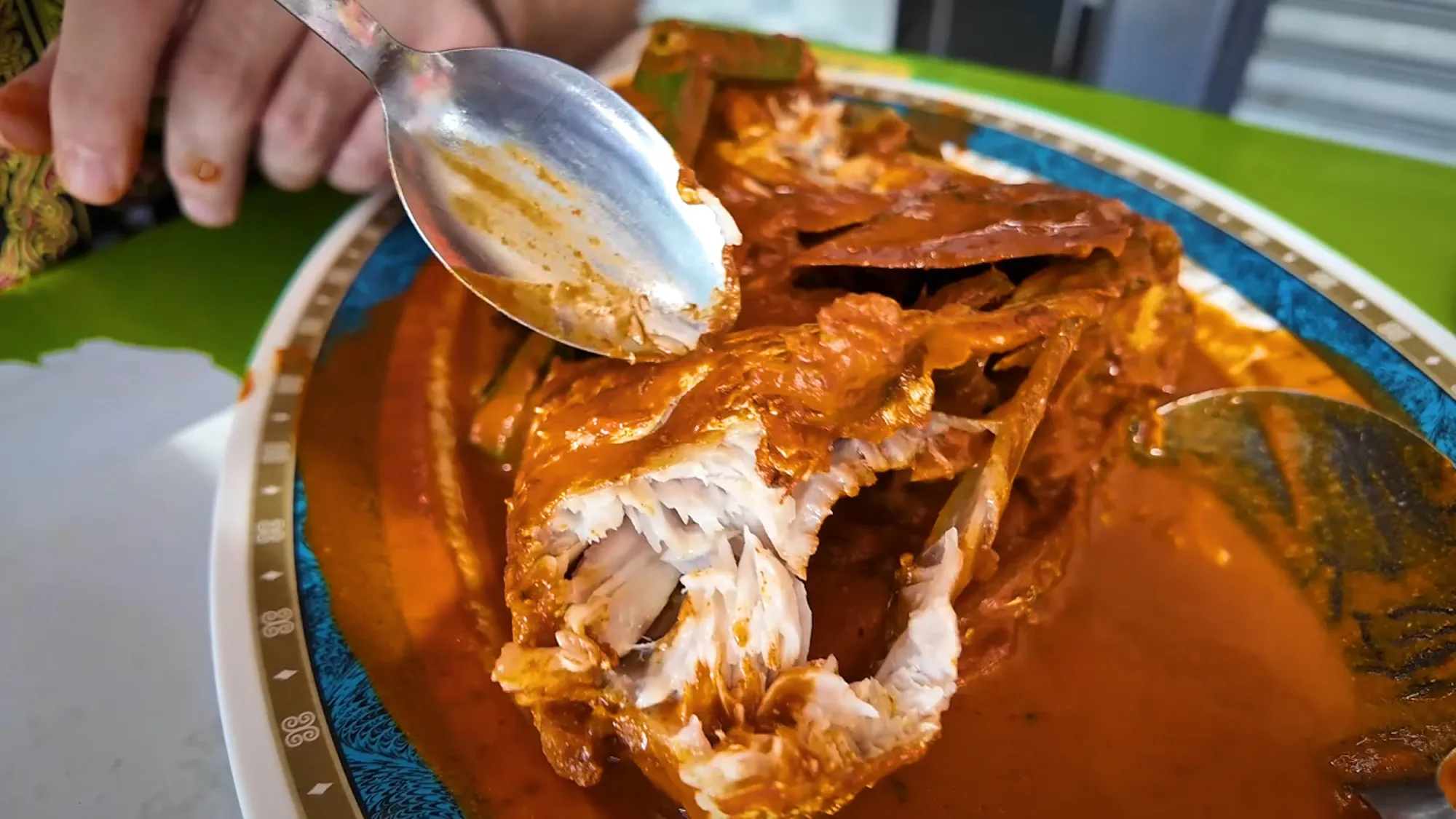 A spoon lifting flaky white meat off a fish head in red curry at Anuar Curry Fish Head in Bangsar, Kuala Lumpur.