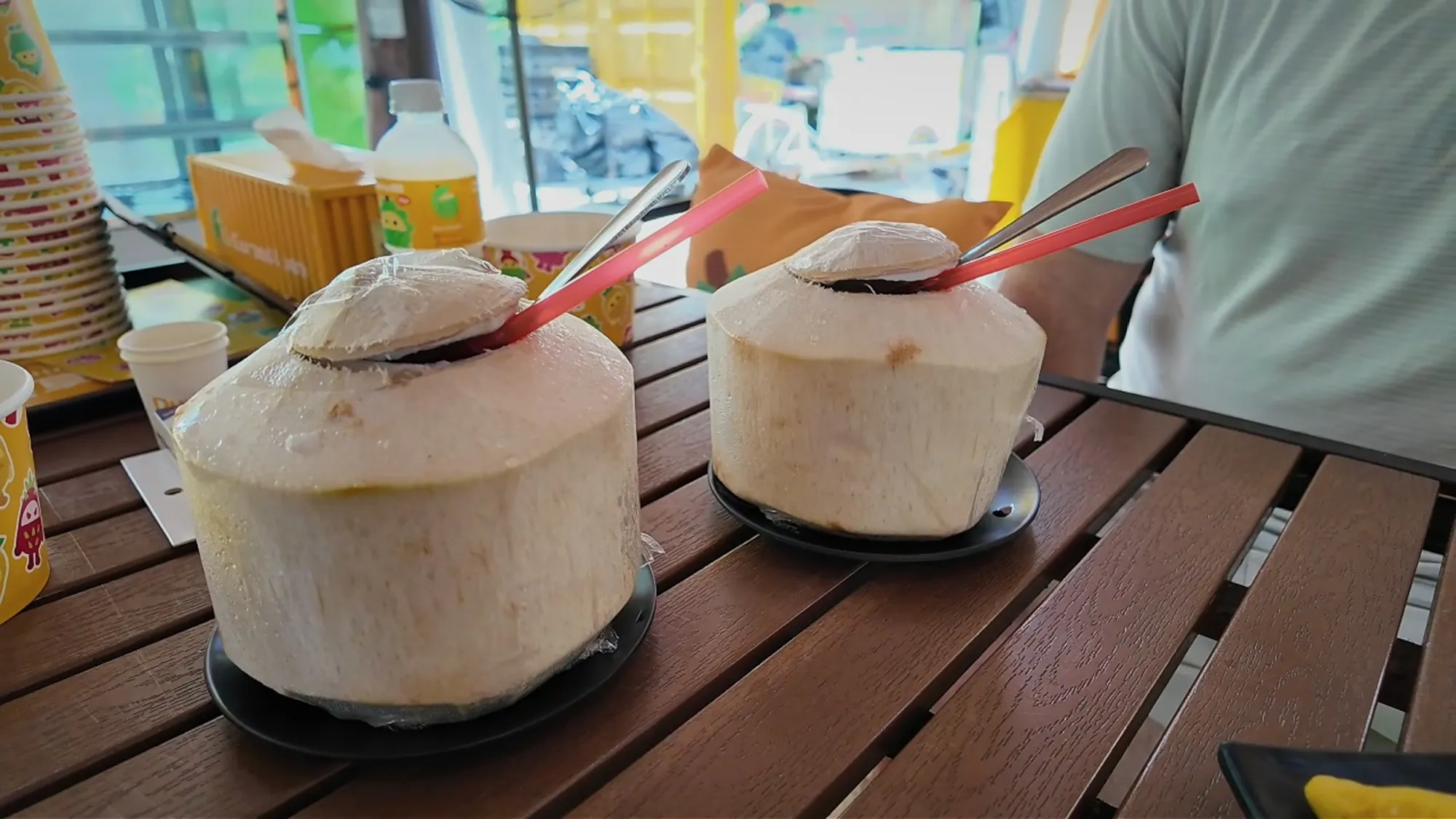 Two fresh young coconuts with straws and spoons on a wooden table, served alongside the durian flight.