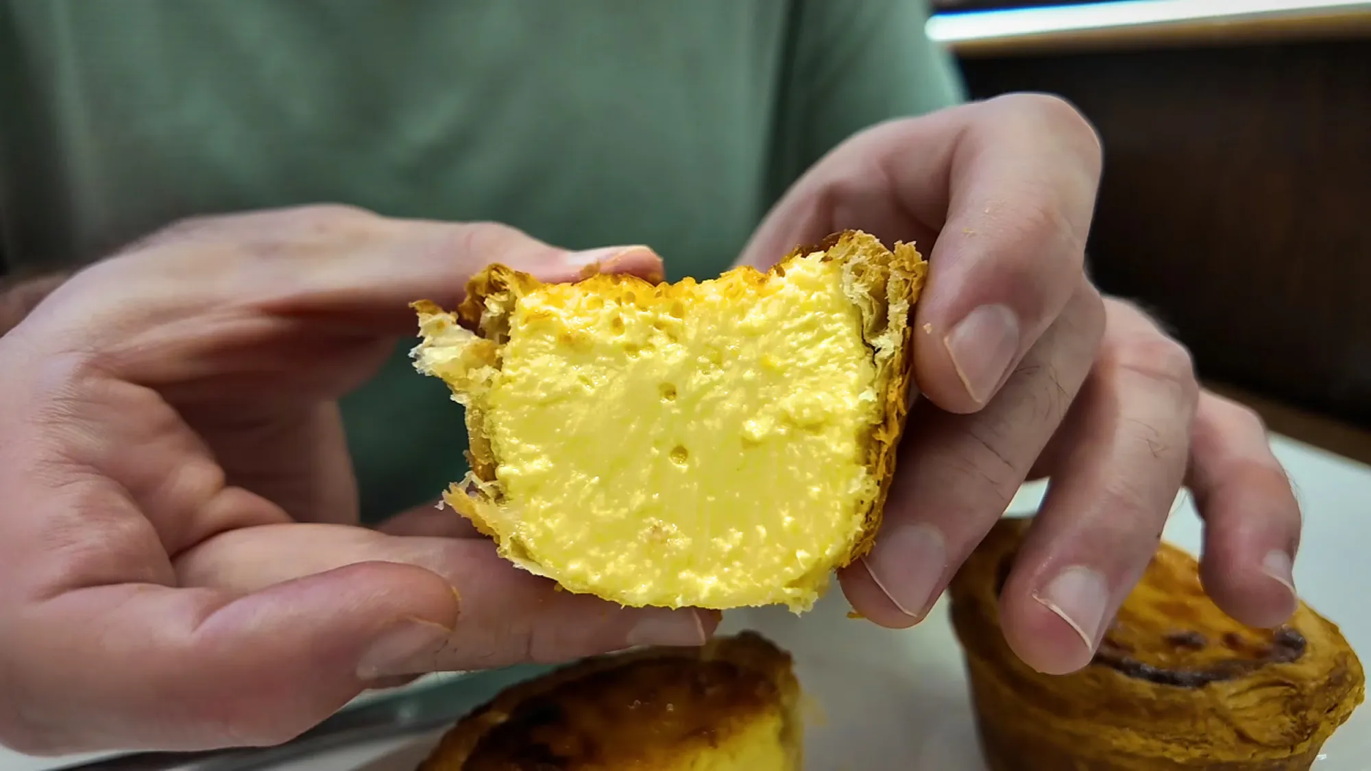 Hands holding an Oriental Kopi egg tart broken in half to show the yellow custard and flaky crust.