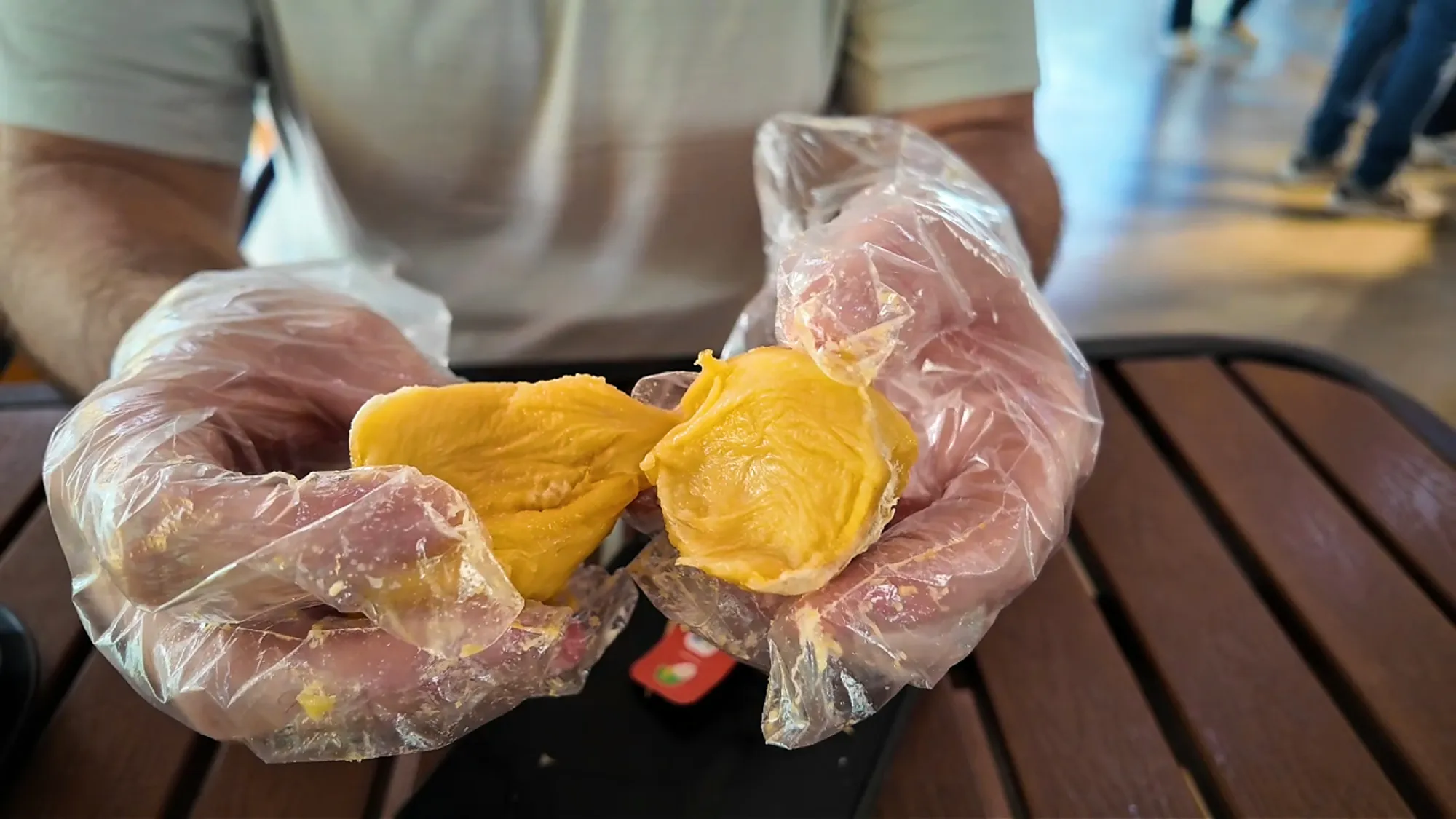 Gloved hands pulling apart a soft, deep yellow piece of durian over a wooden table.