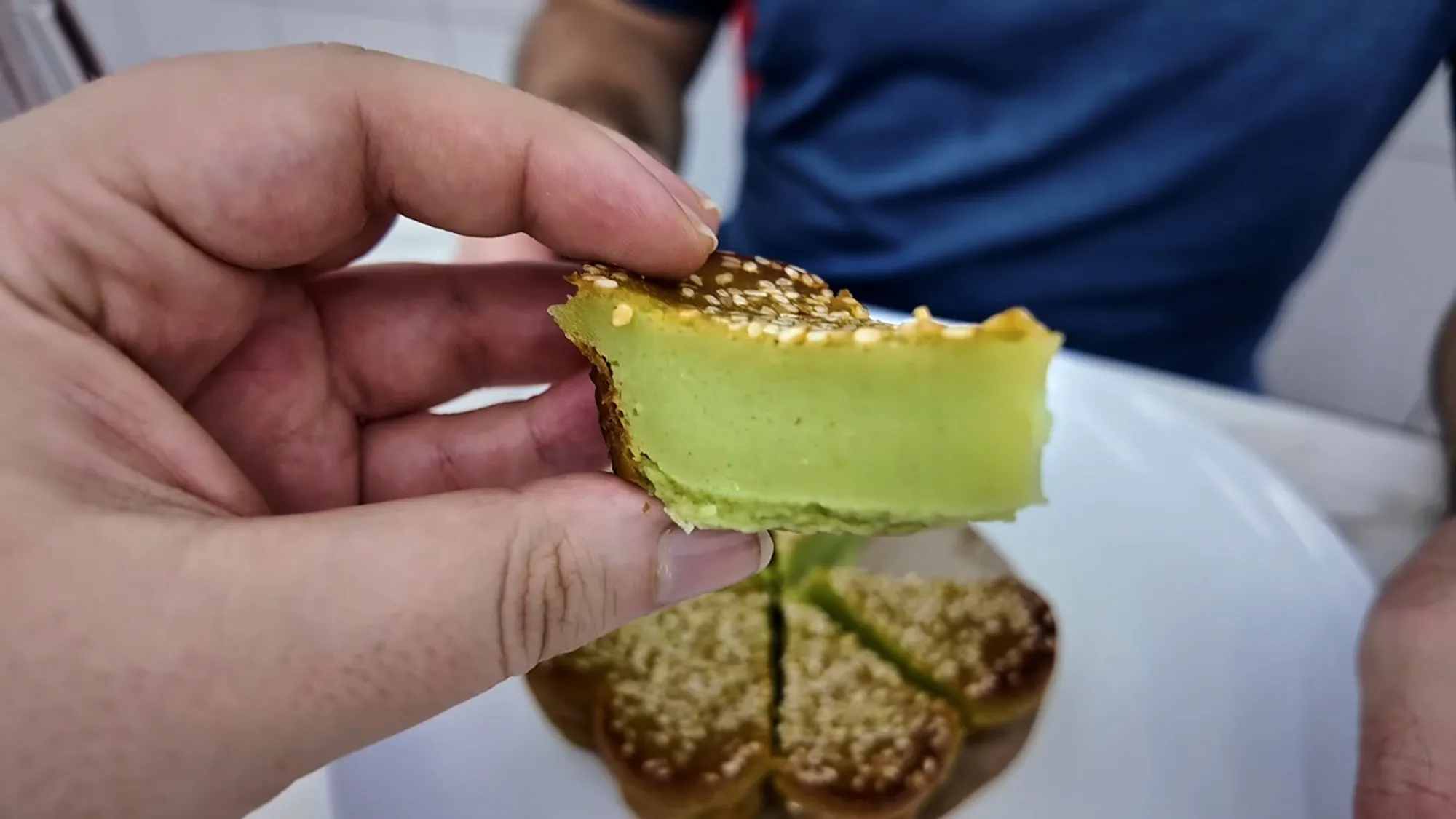 A hand holding up a slice of kuih bakar pandan to show its soft green inside, above the sesame-topped slices on the plate.