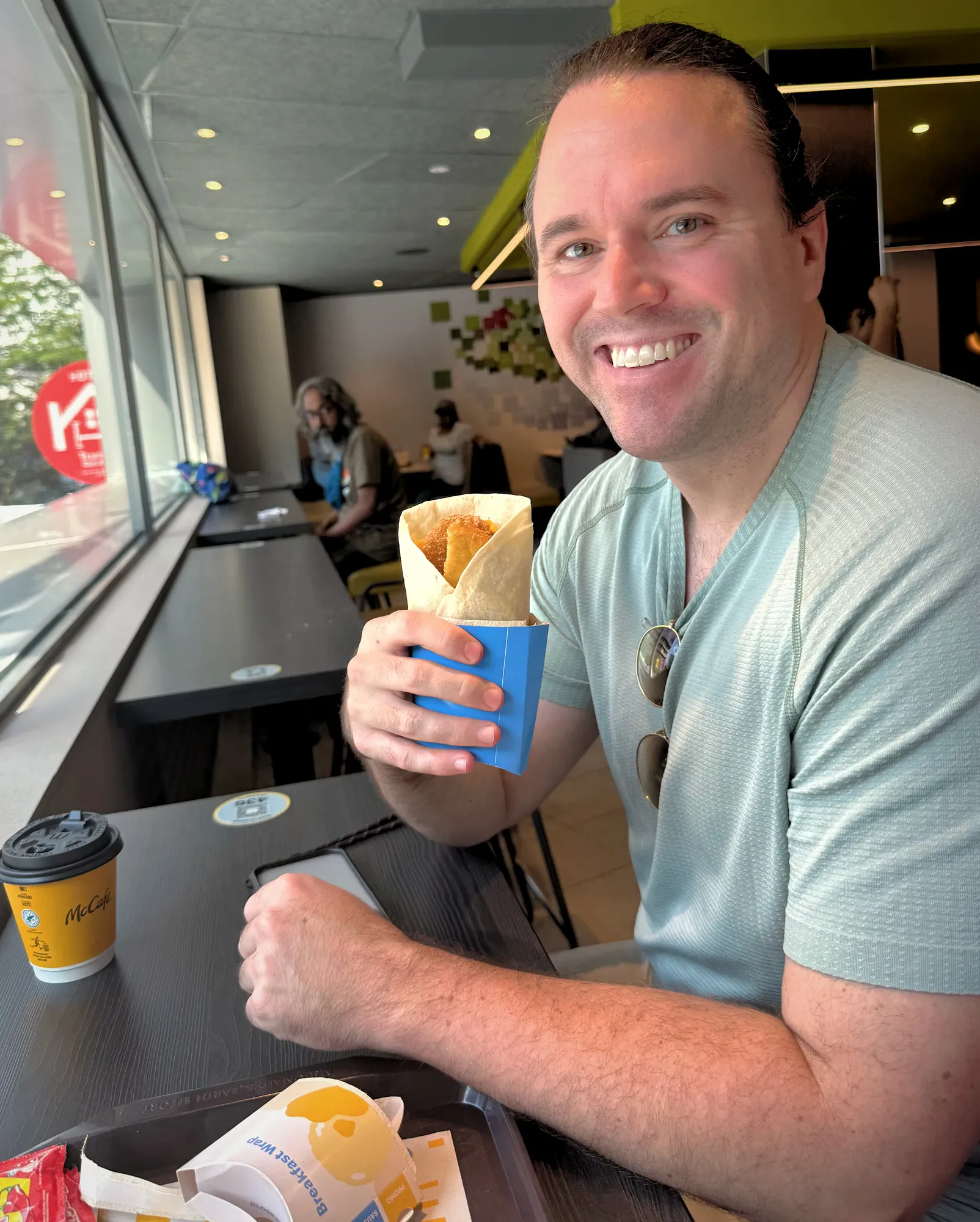 A smiling man sits by a sunny cafe window holding a McDonald's breakfast wrap with a hash brown inside.
