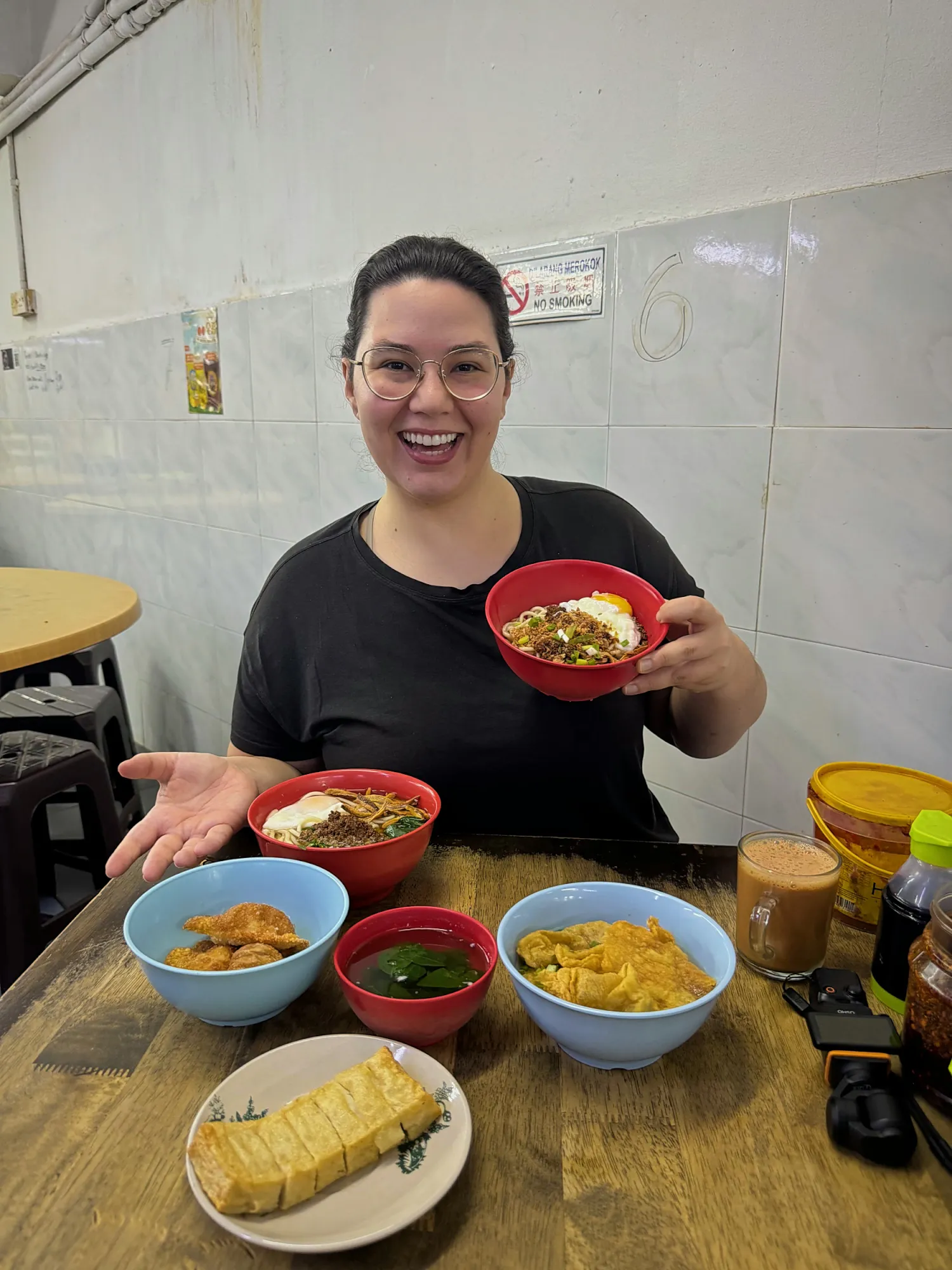 Bernie smiling and holding up a bowl of chili pan mee over our spread of sides at Restoran Kin Kin in Kuala Lumpur.