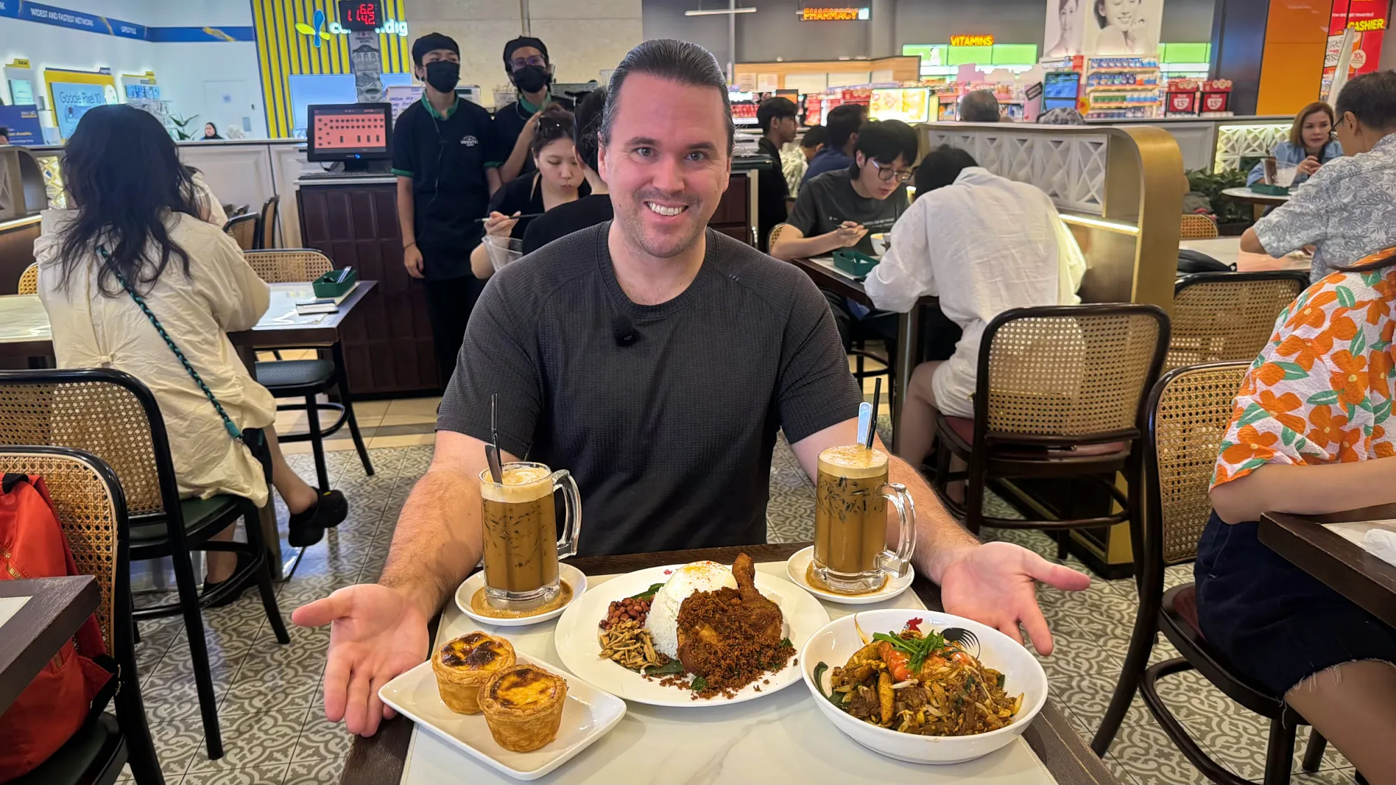 A smiling man at a table with curry laksa, egg tarts, a polo bun, kaya toast, and iced drinks at Oriental Kopi.