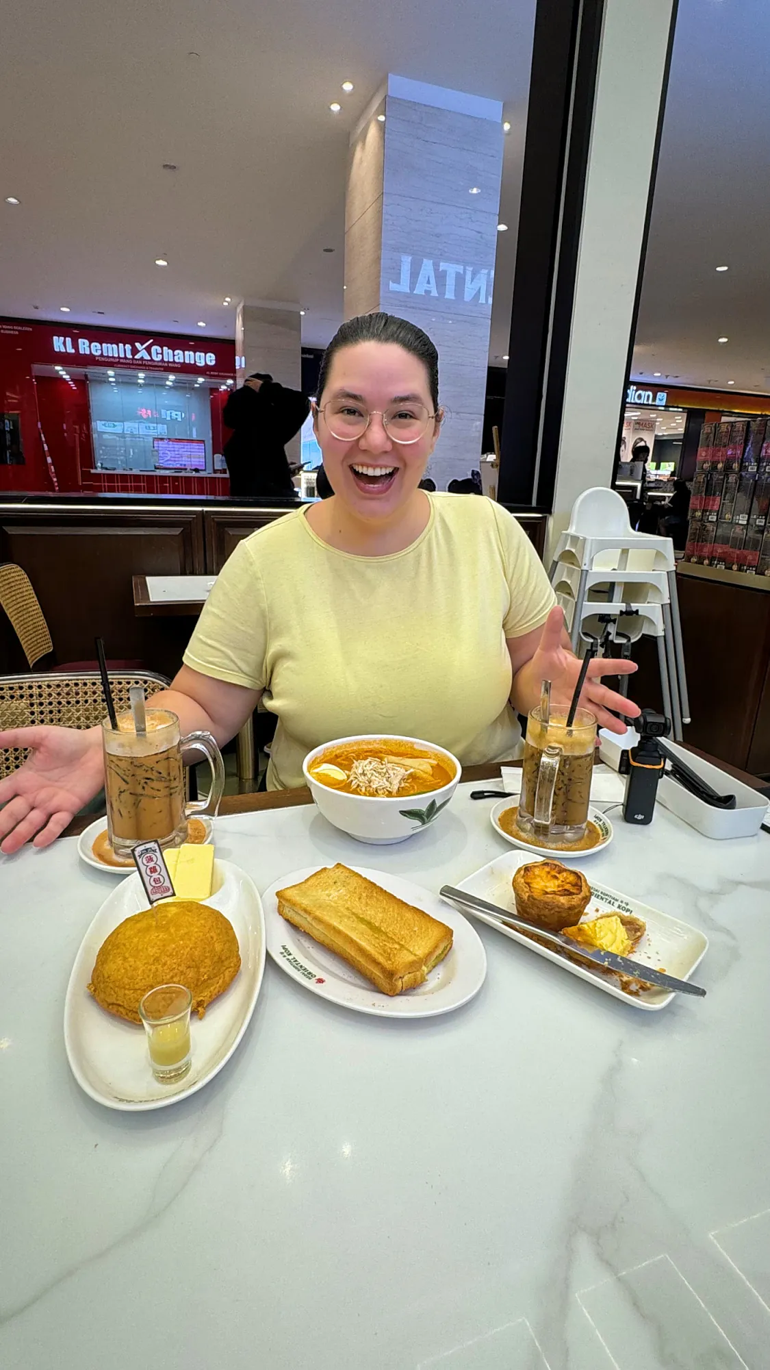 A smiling woman holding a plate of egg tarts with two iced drinks at Oriental Kopi.