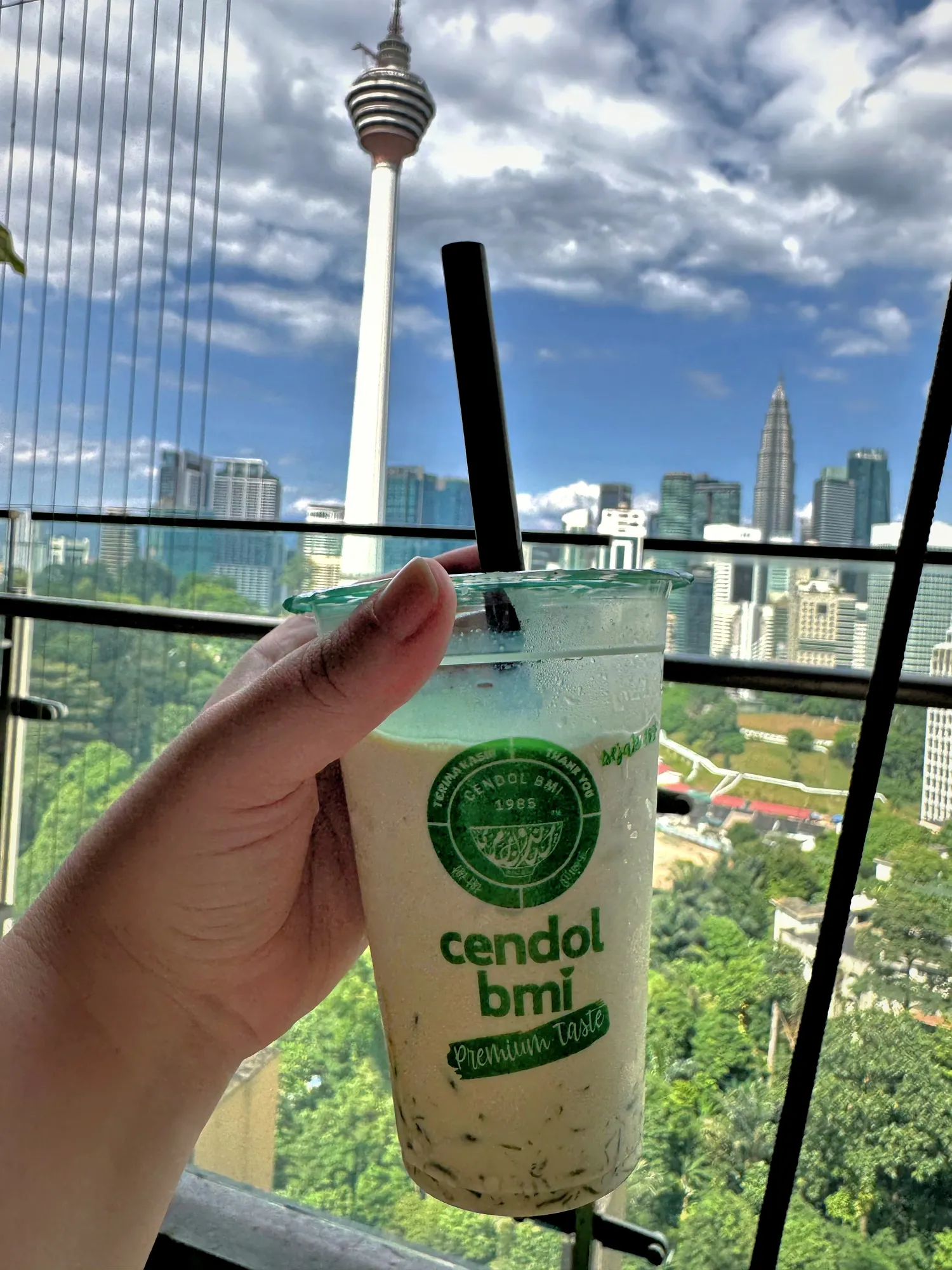 Holding a tall cup of iced cendol against a sunny view of the KL Tower