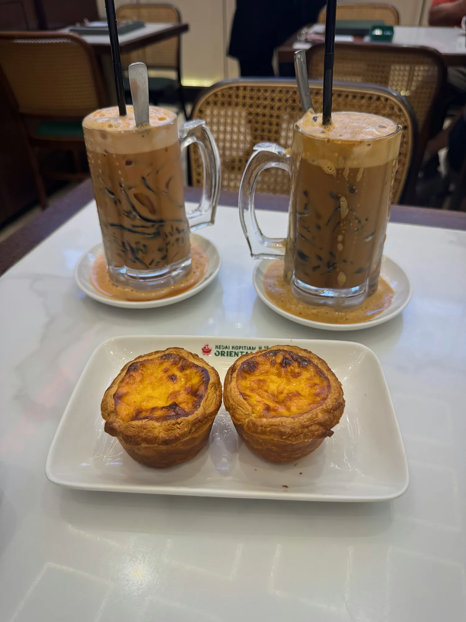 Two tall mugs of iced milk tea next to a plate of two egg tarts at Oriental Kopi.