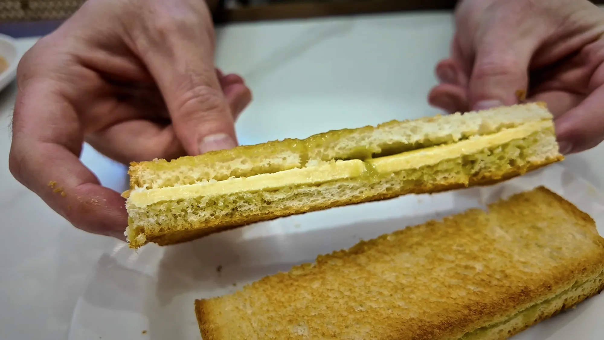 Hands holding a slice of Oriental Kopi kaya butter toast showing green pandan kaya and thick butter inside.
