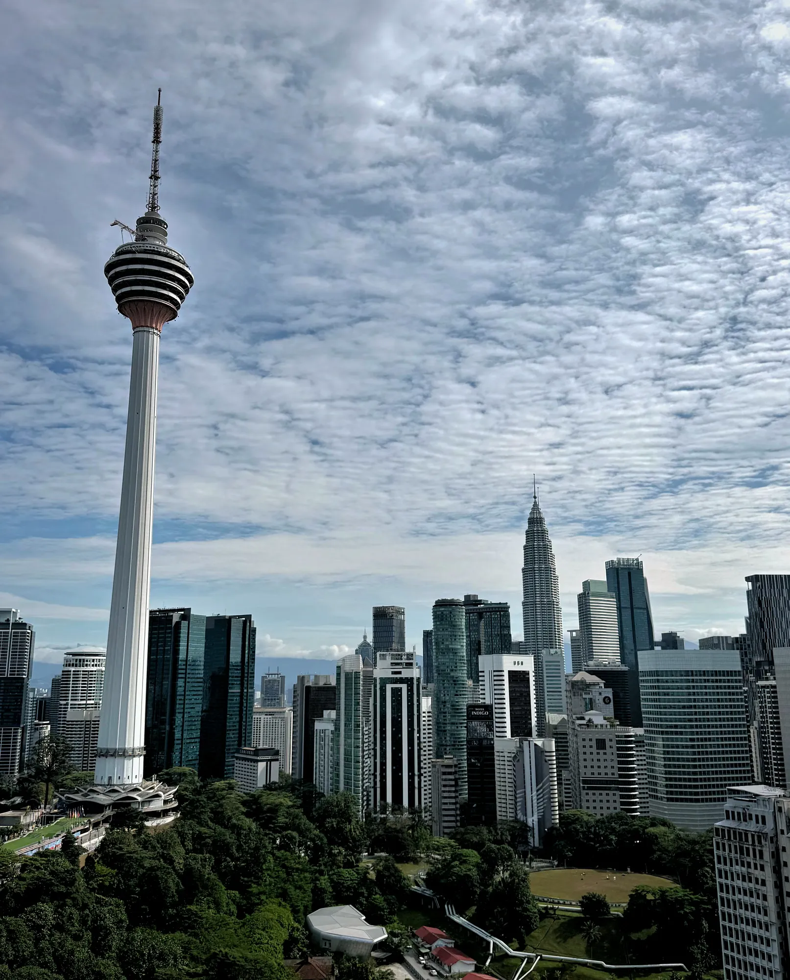 The white KL Tower against a partly cloudy sky, with the Petronas Towers and the Kuala Lumpur skyline clearly visible behind it.