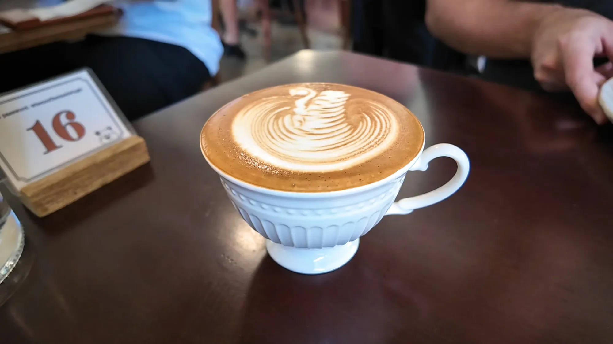 Toffee nut latte with swan latte art in a white ribbed cup, filled to the brim, next to a table number stand.